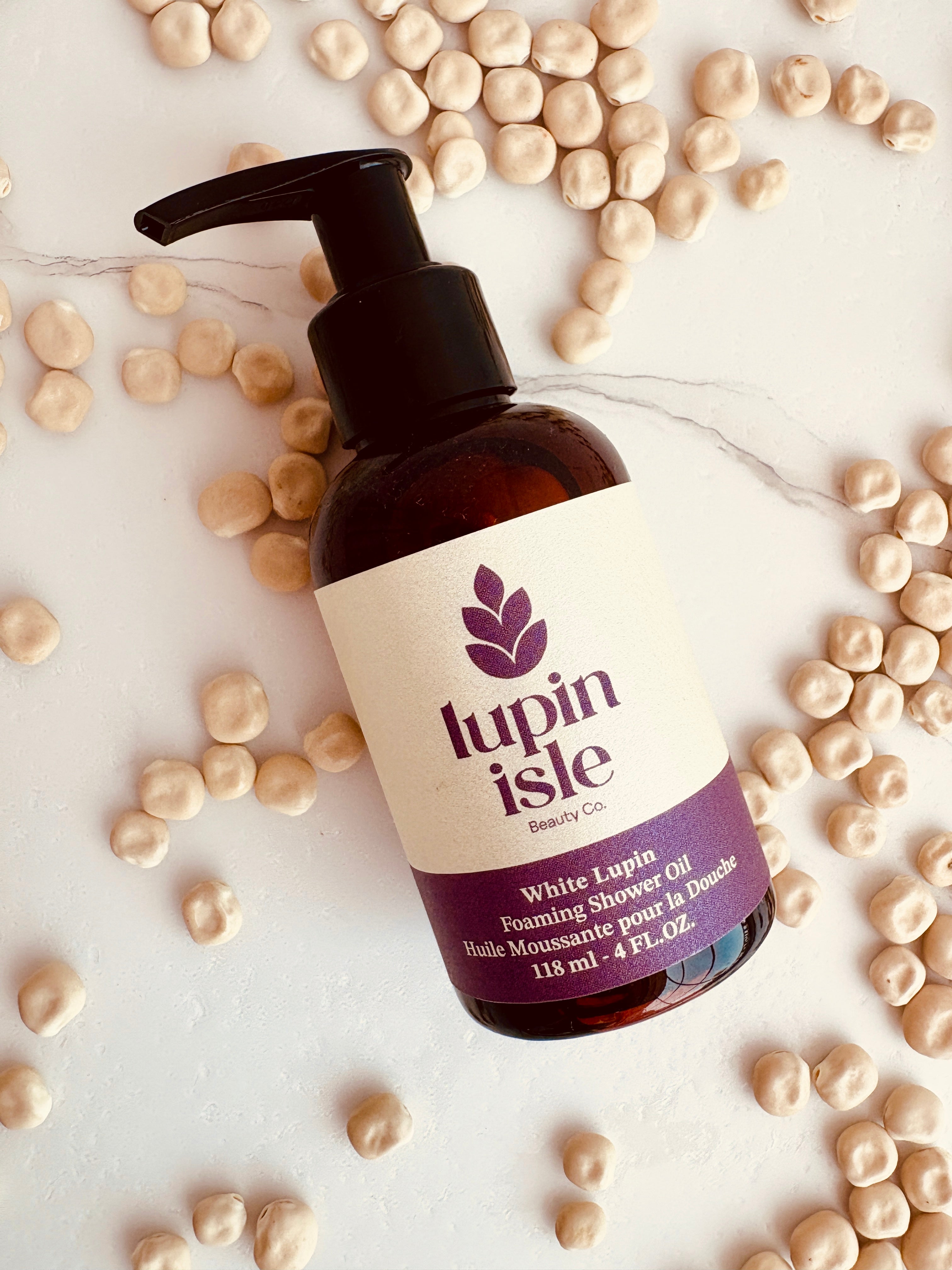 A bottle of Lupin Isle Beauty Co. White Lupin Foaming Shower Oil, styled on a white marble surface surrounded by scattered white lupin seeds.