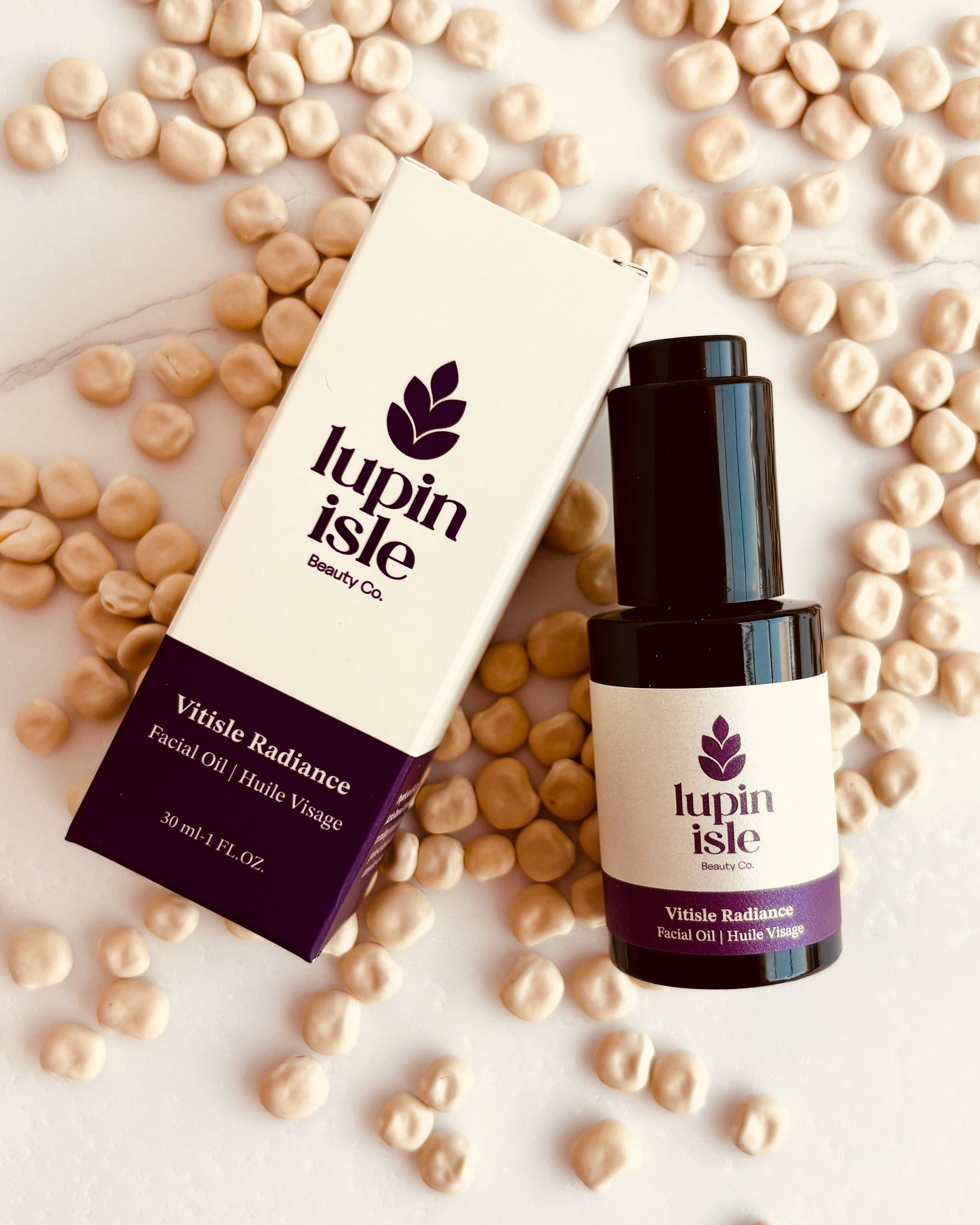 A bottle of Lupin Isle Vitale Radiance Facial Oil next to its white and dark purple packaging box, styled on a white marble surface surrounded by scattered lupin beans.