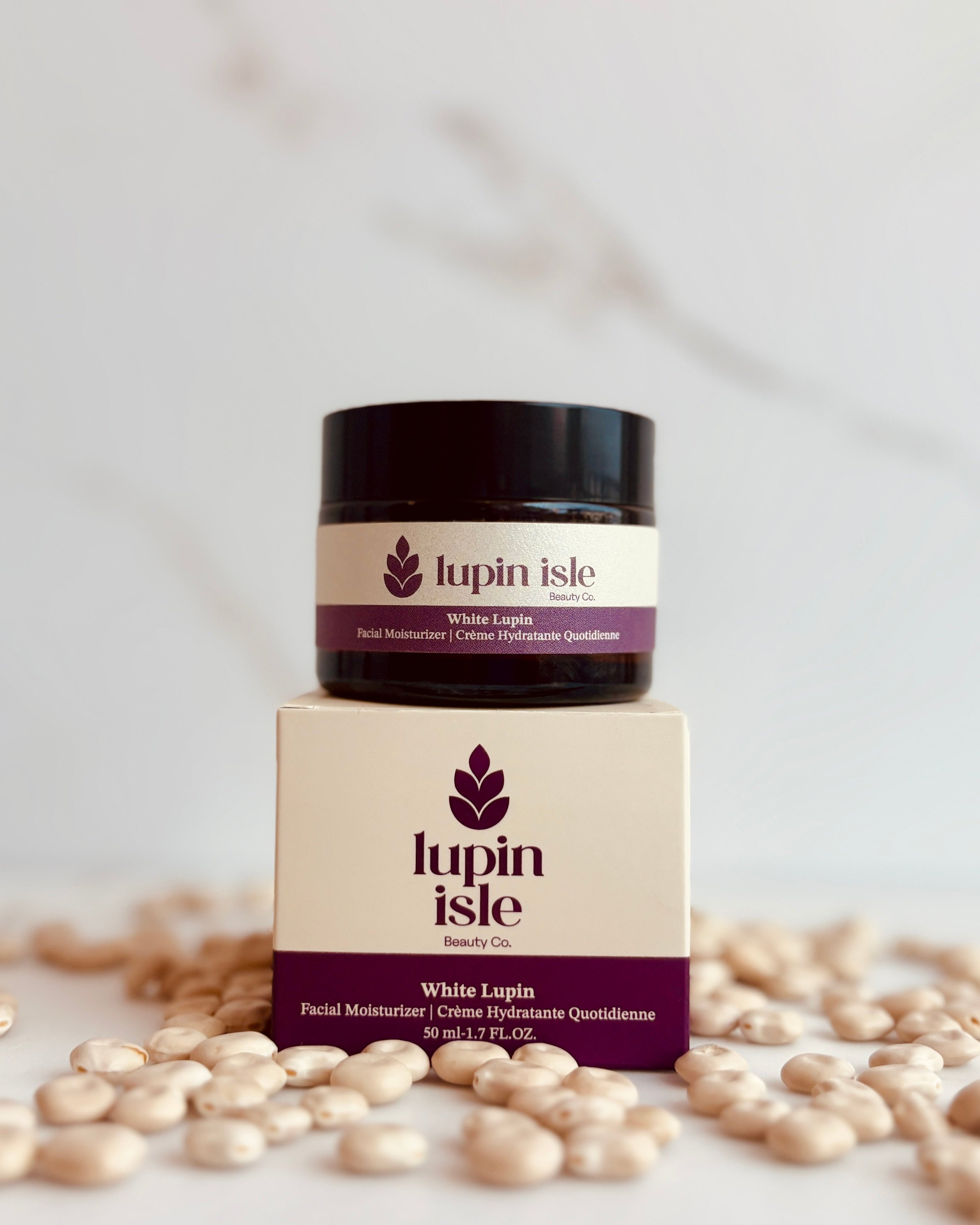A small jar of lupin Isle White Lupin facial moisturizer sits on top of its branded box, surrounded by scattered lupin beans on a light marble-like surface.