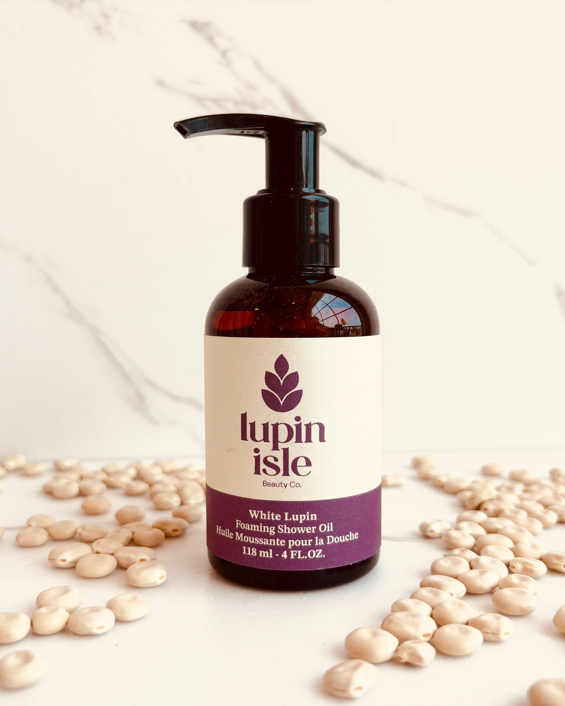 A bottle of Lupin Isle White Clover Steaming Shower Oil with a black pump dispenser, set on a white marble surface surrounded by scattered white lupin beans.