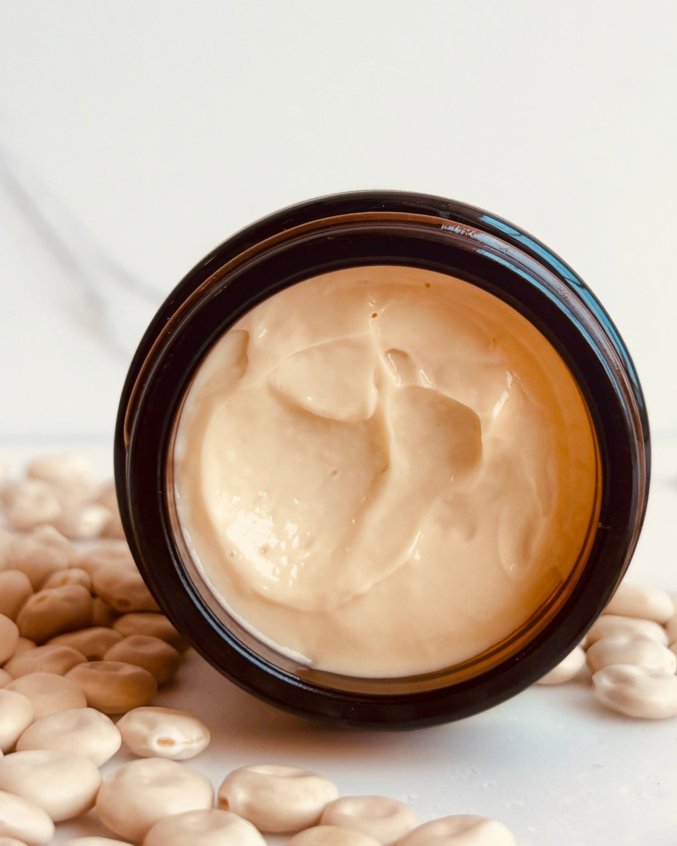 An open dark amber cosmetic jar filled with a thick beige cream, surrounded by scattered lupin beans on a light marble-like surface.