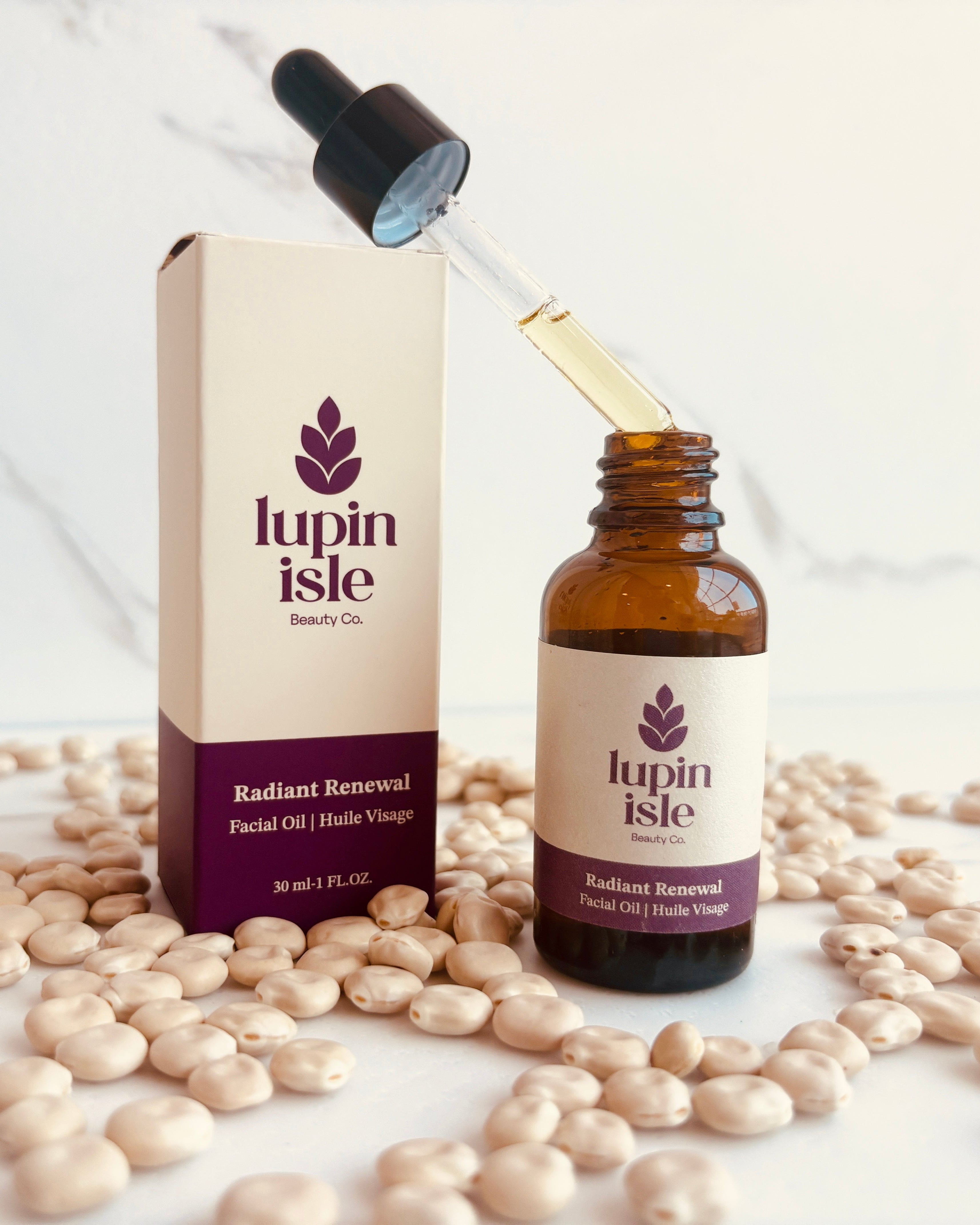 Lupin Isle Radiant Renewal Facial oil shown with its amber glass dropper bottle and matching cream-colored box, surrounded by scattered lupin beans on a light marble surface.