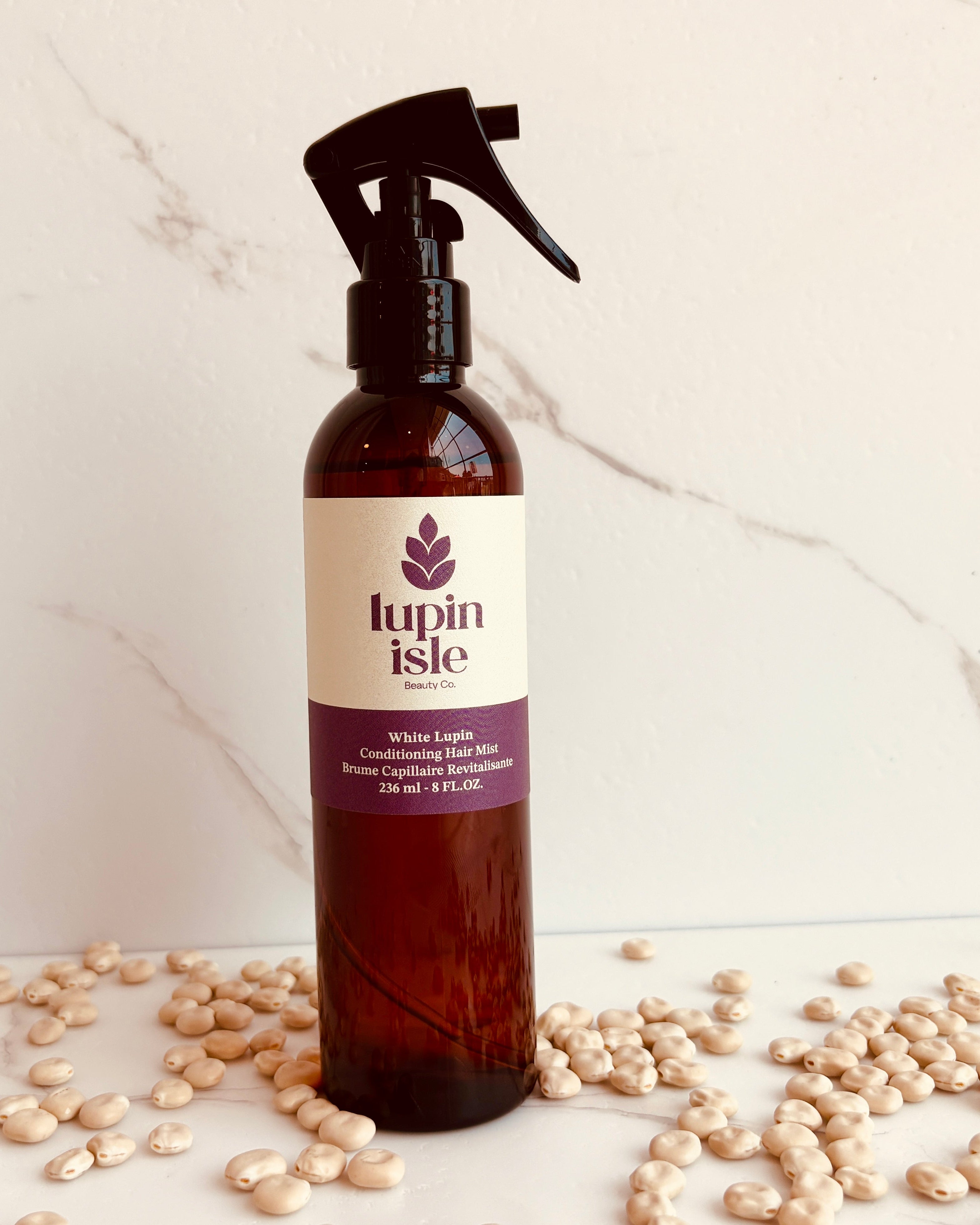 A bottle of Lupin Isle Conditioning Hair mist with a black spray top and cream label, standing on a marble surface surrounded by scattered lupin beans.