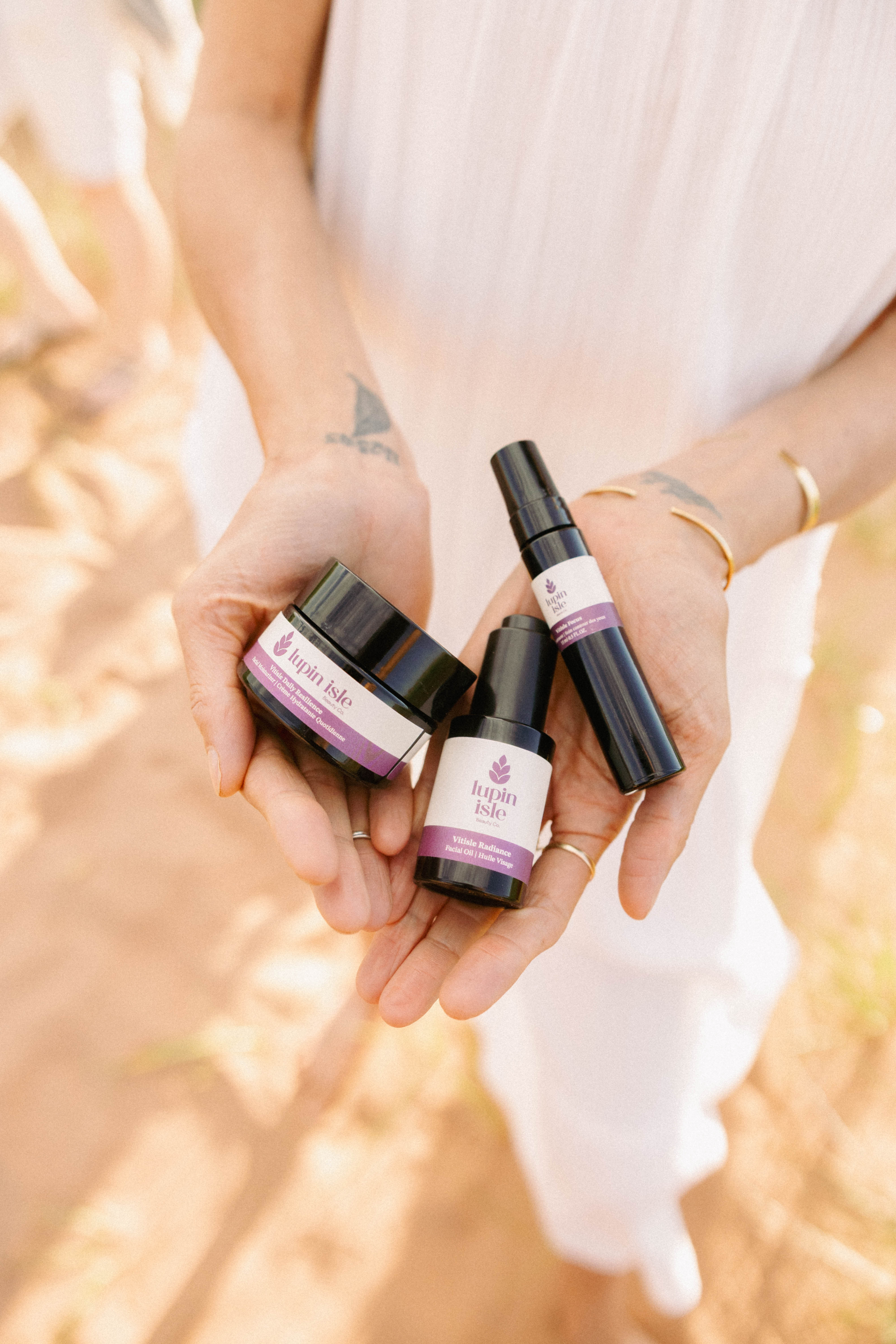 A person in a white dress holds three small wellness products in their hands outdoors. The products include a black jar, a black spray bottle, and a black dropper bottle with purple and white labels.
