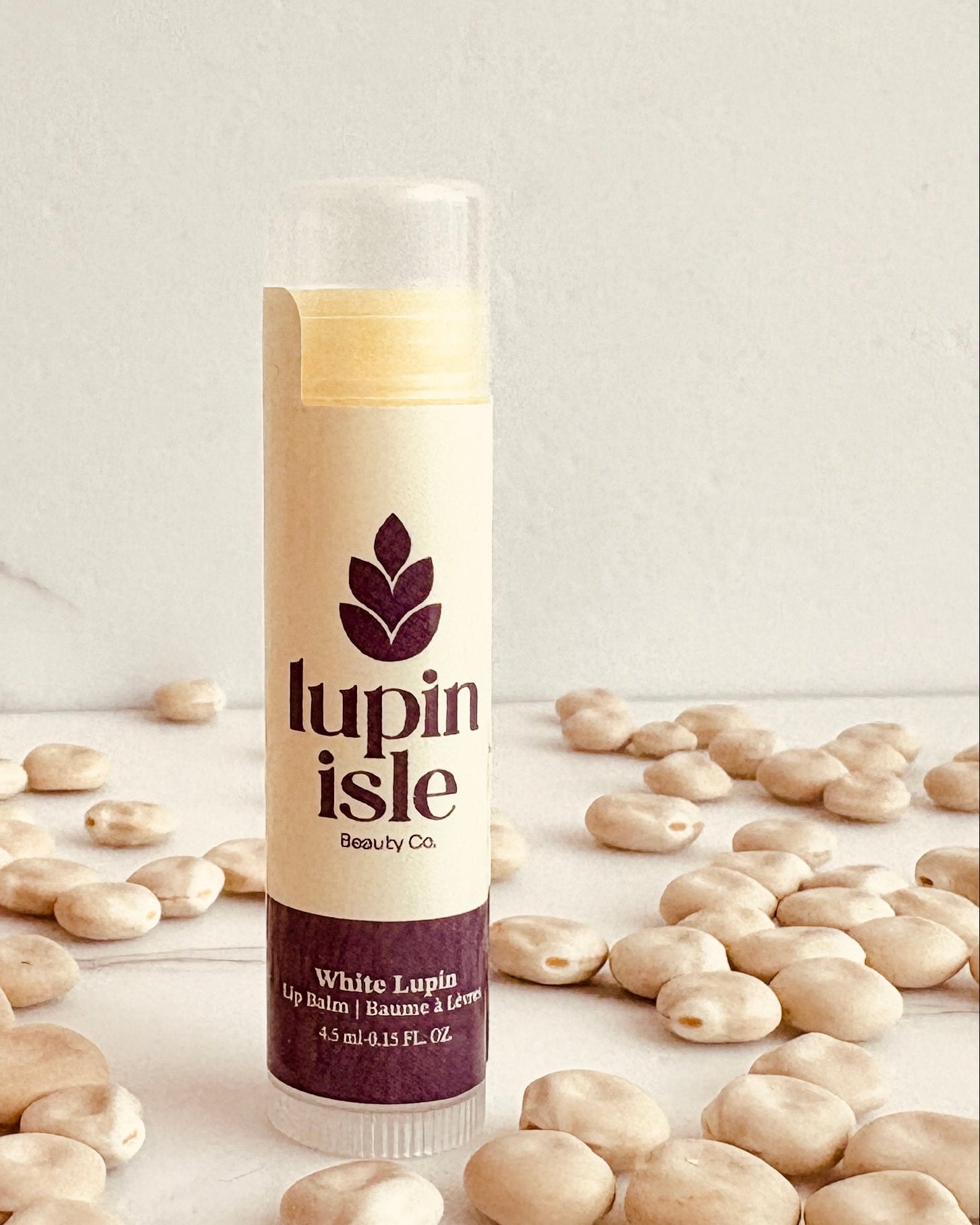 A tube of Lupin Isle White Lupin lip balm standing upright on a marble-like surface surrounded by scattered white lupin beans. The product packaging is cream with a dark purple brand logo and text, and the background is a light marble wall.