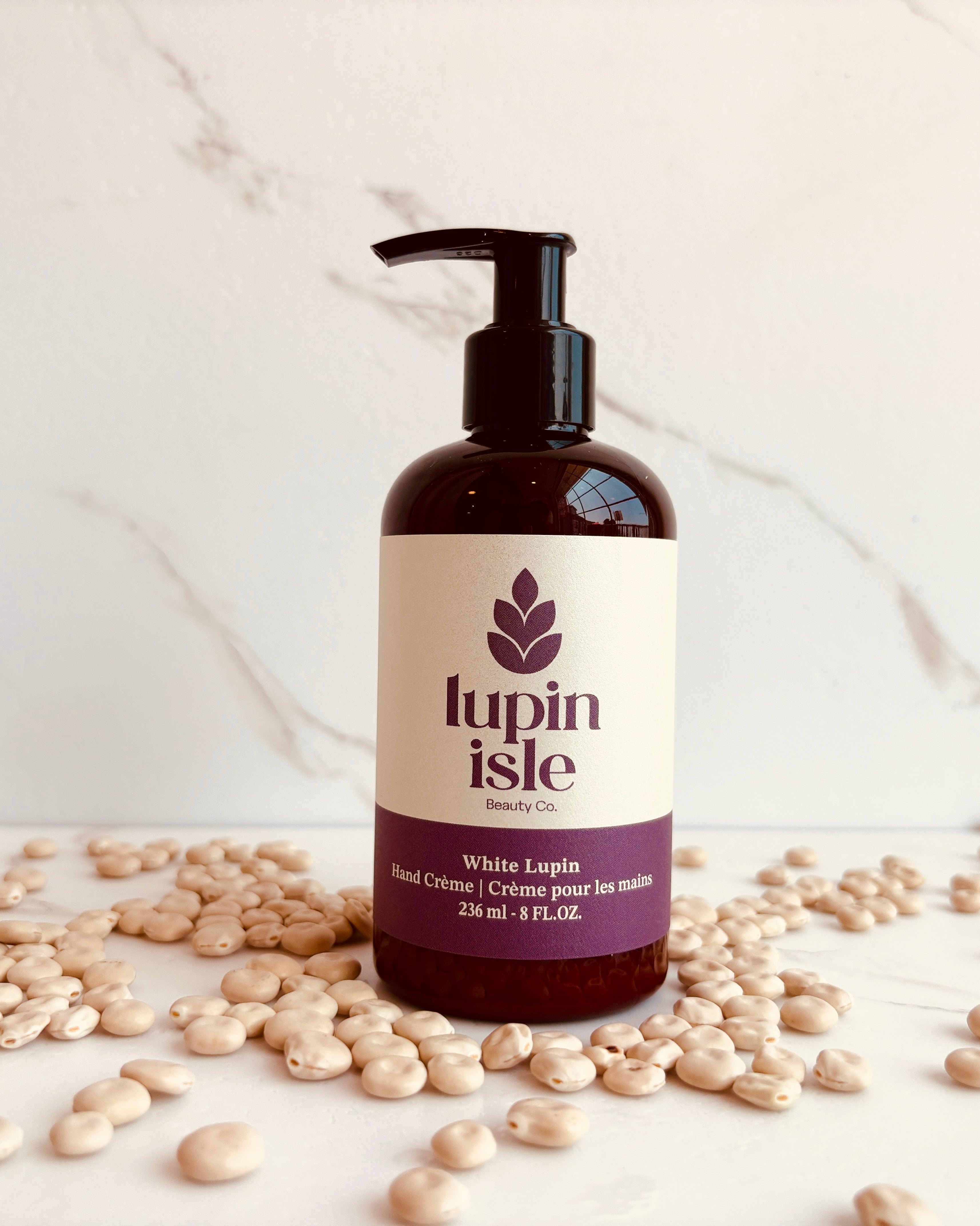 A bottle of Lupin Isle White Lupin Hand Cream with a black pump dispenser, set on a white marble surface scattered with white lupin beans.