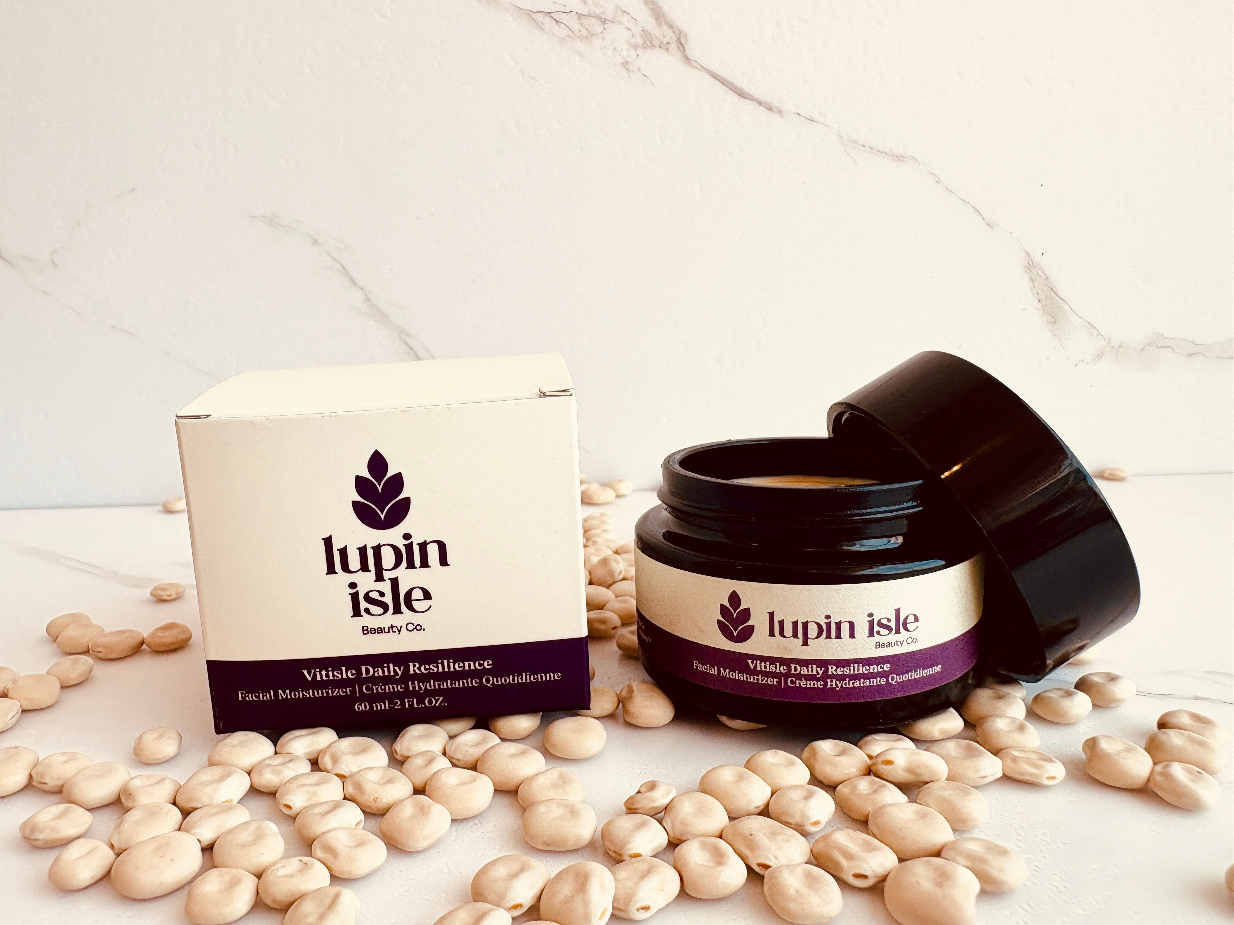 A jar of Lupin Isle Vitalize Daily Resilience facial moisturizer next to its white and purple packaging box, styled with dried beans on a white marble surface.
