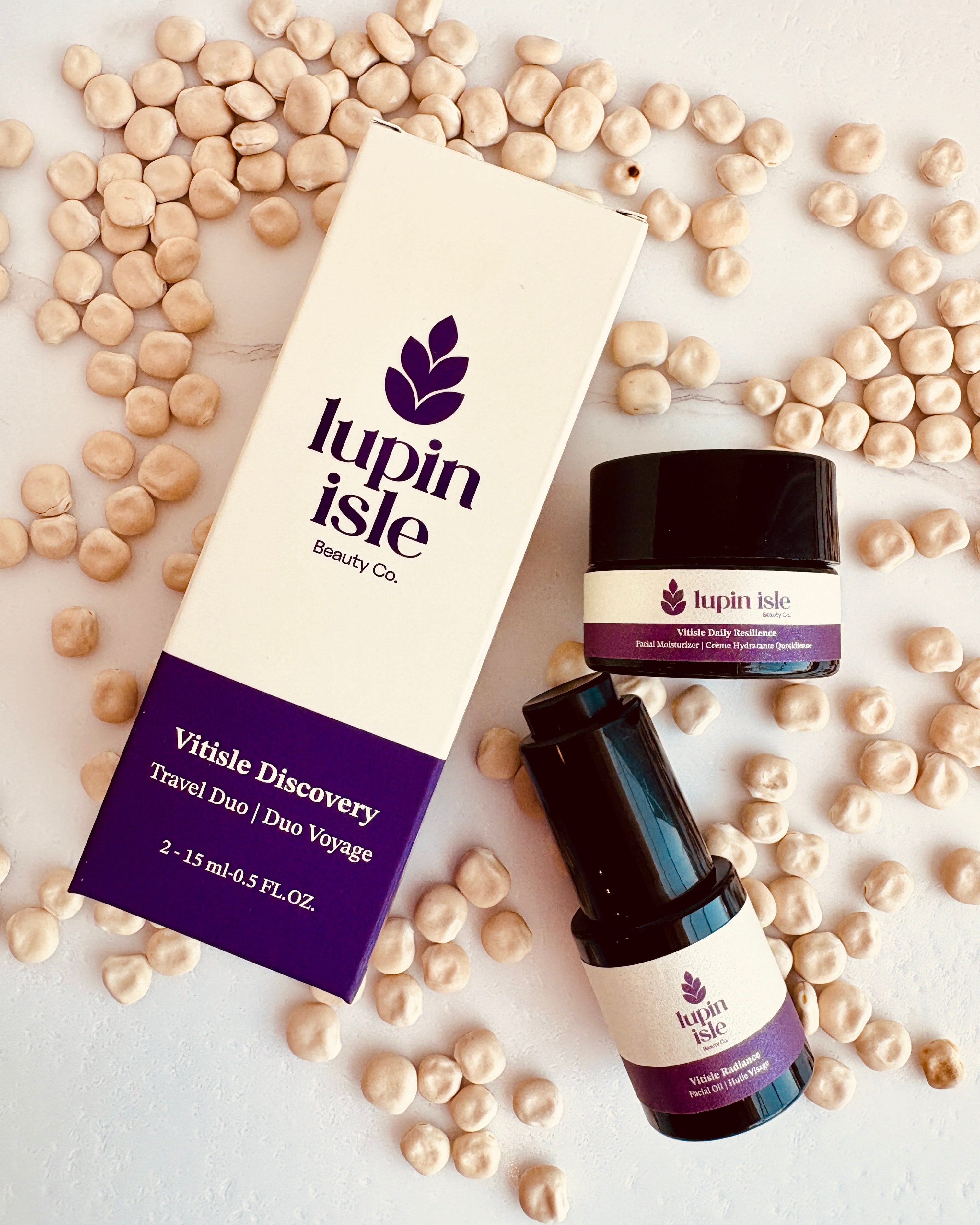 Lupin Isle Vitalie Discovery Travel Duo set, featuring a cream and purple cardboard box, a small jar, and a dark amber bottle, arranged on a white surface with scattered lupin beans.
