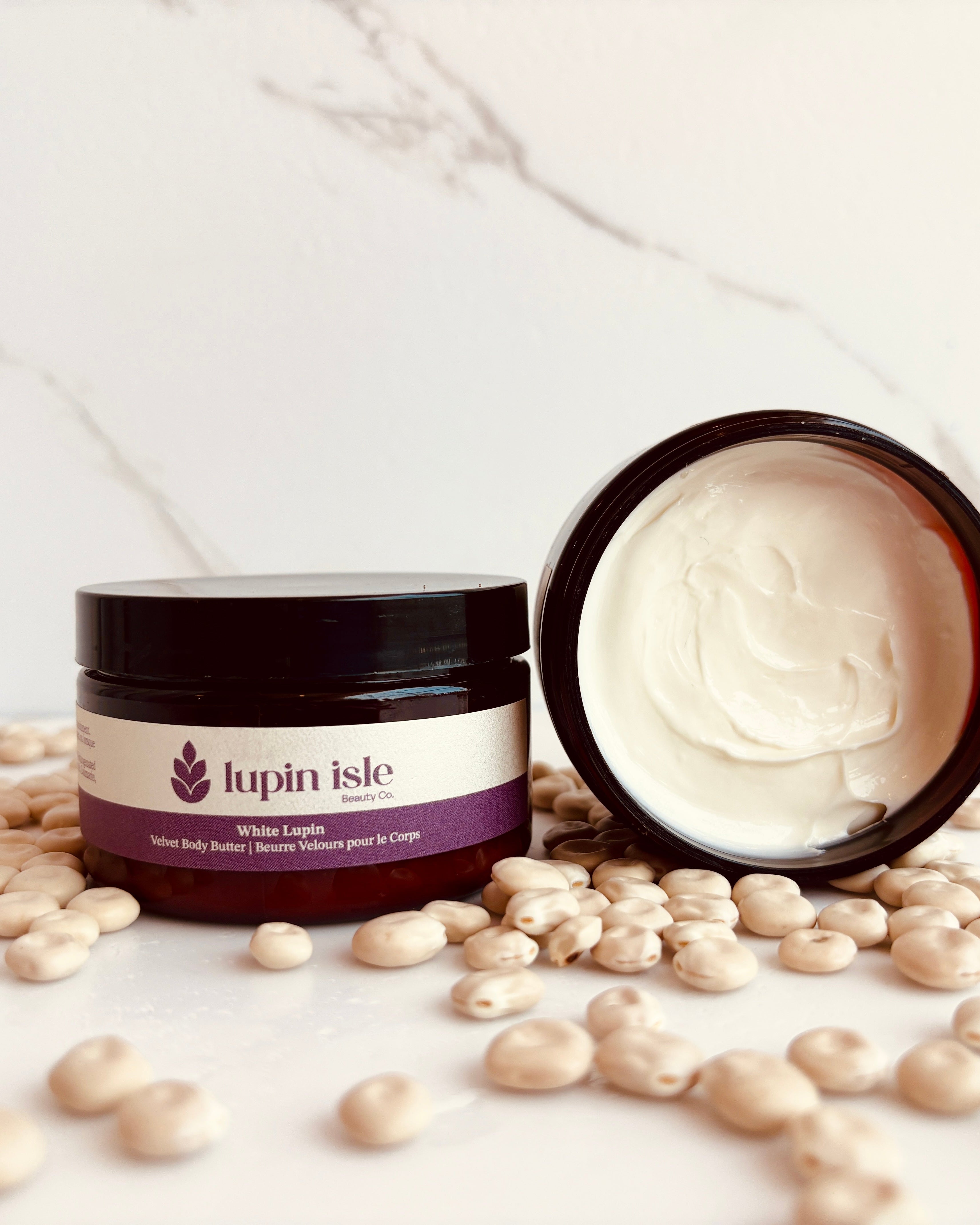 A jar of Lupin Isle Velvet Body Butter, with one jar standing upright and another lying on its side to show the creamy texture inside. The jars are surrounded by scattered lupin seeds on a white marble surface and background.