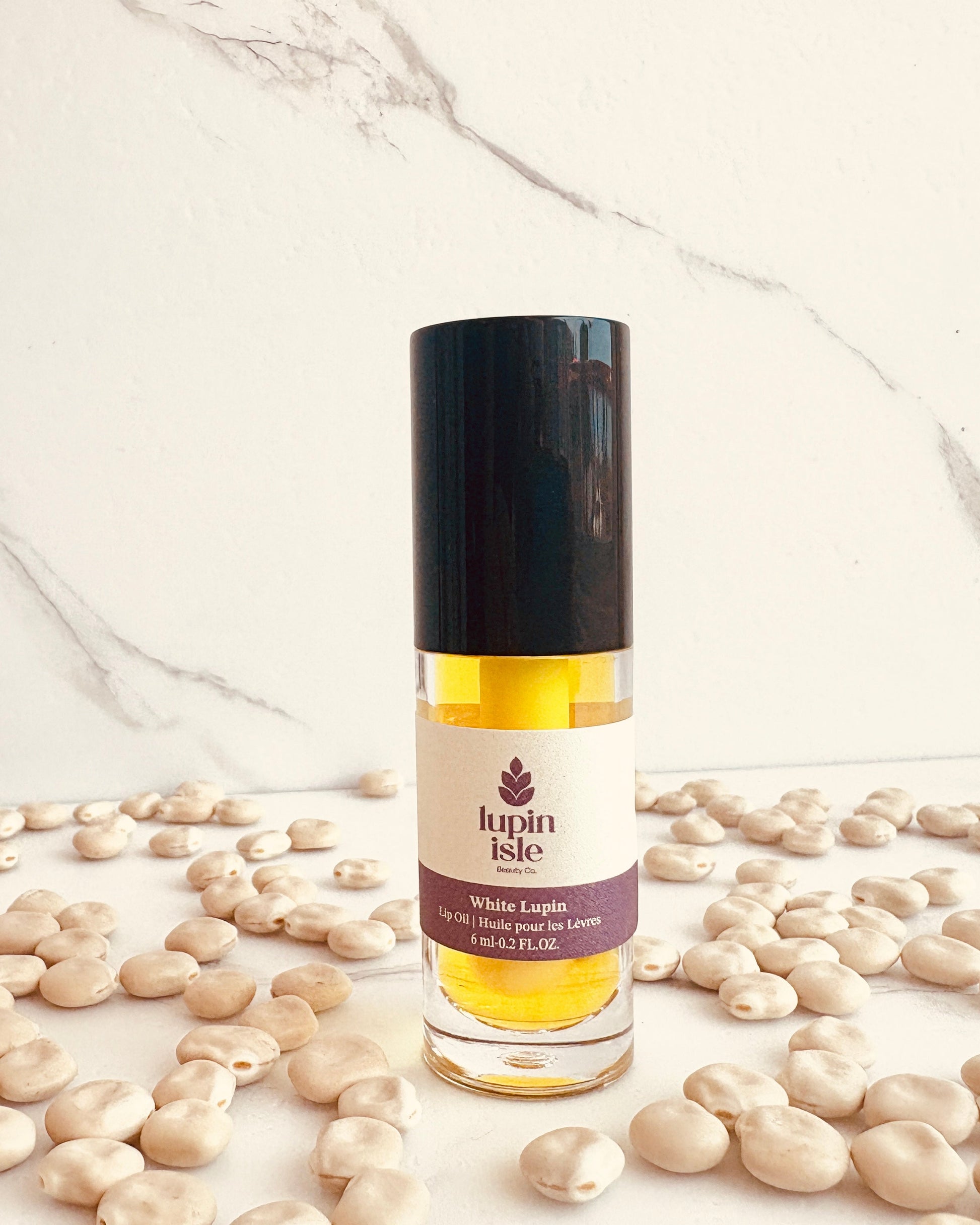 A bottle of Lupin Isle White Lupin lip oil with a black cap, surrounded by scattered beige lupin seeds on a white marble background.