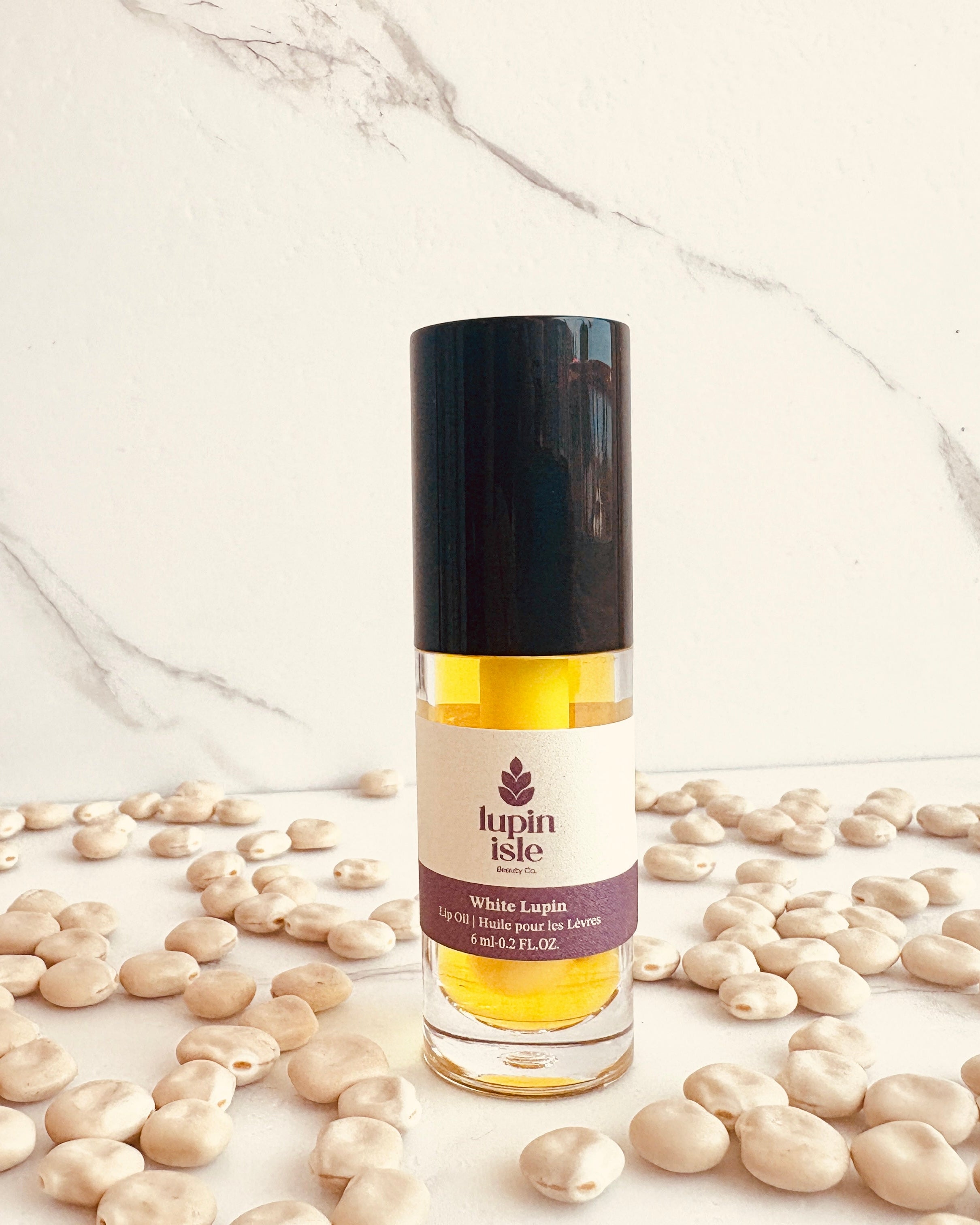 A bottle of Lupin Isle White Lupin lip oil with a black cap, surrounded by scattered beige lupin seeds on a white marble background.