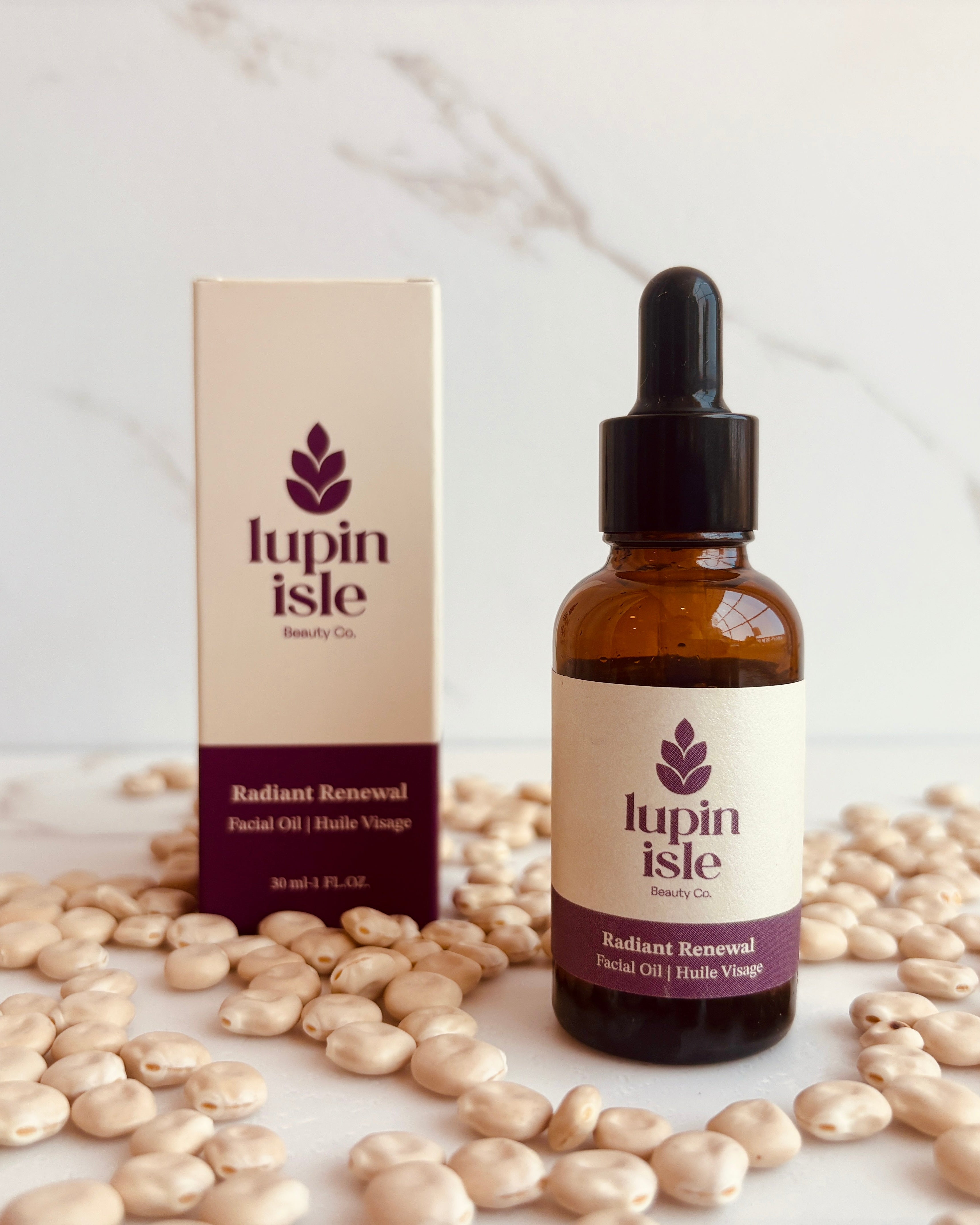 A bottle of Lupin Isle Radiant Renewal Facial Oil next to its packaging box, styled on a white marble surface surrounded by scattered lupin beans.