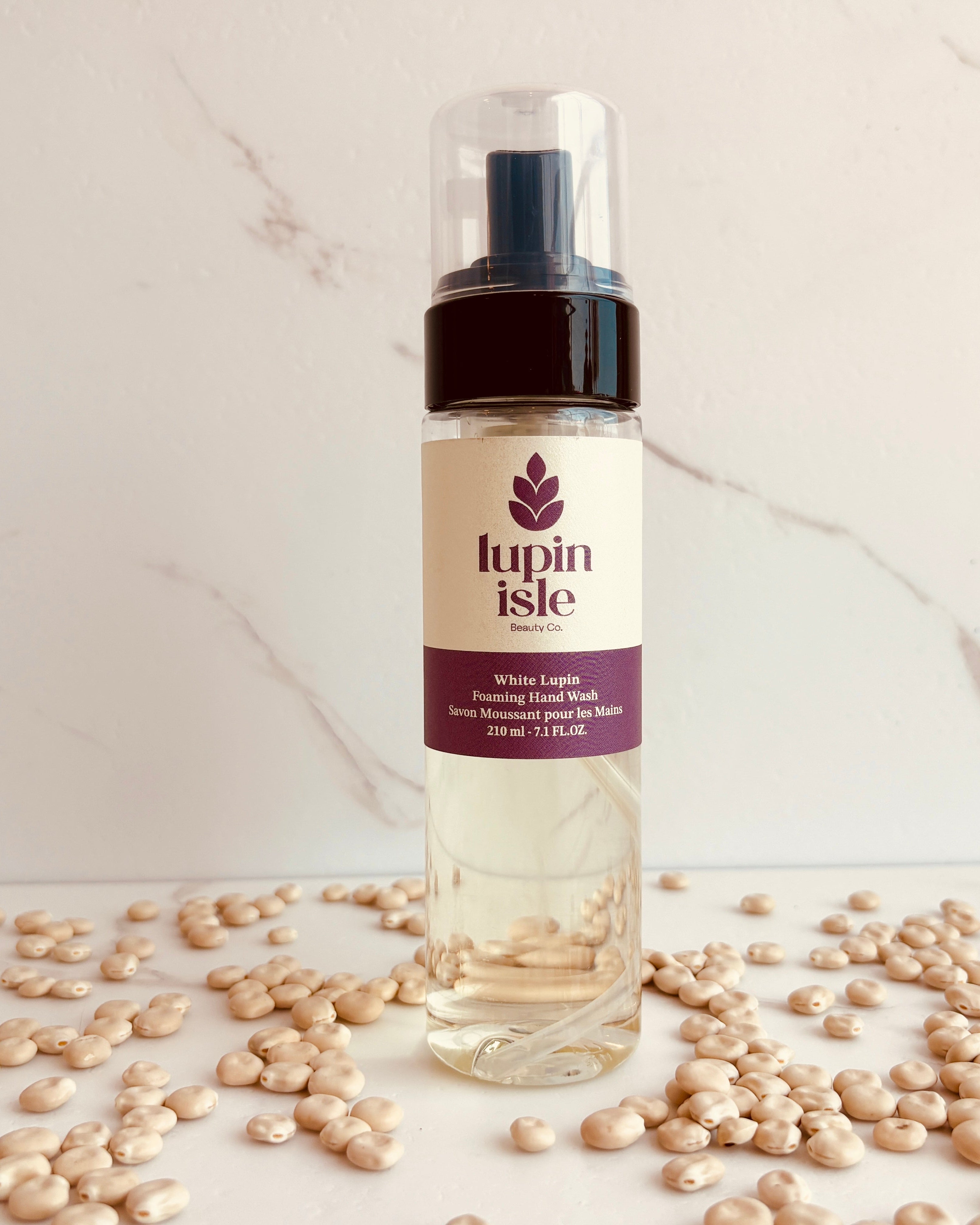A bottle of Lupin Isle White Lupin Hair Foaming Hand Wash standing on a marble-like surface scattered with white lupin beans.