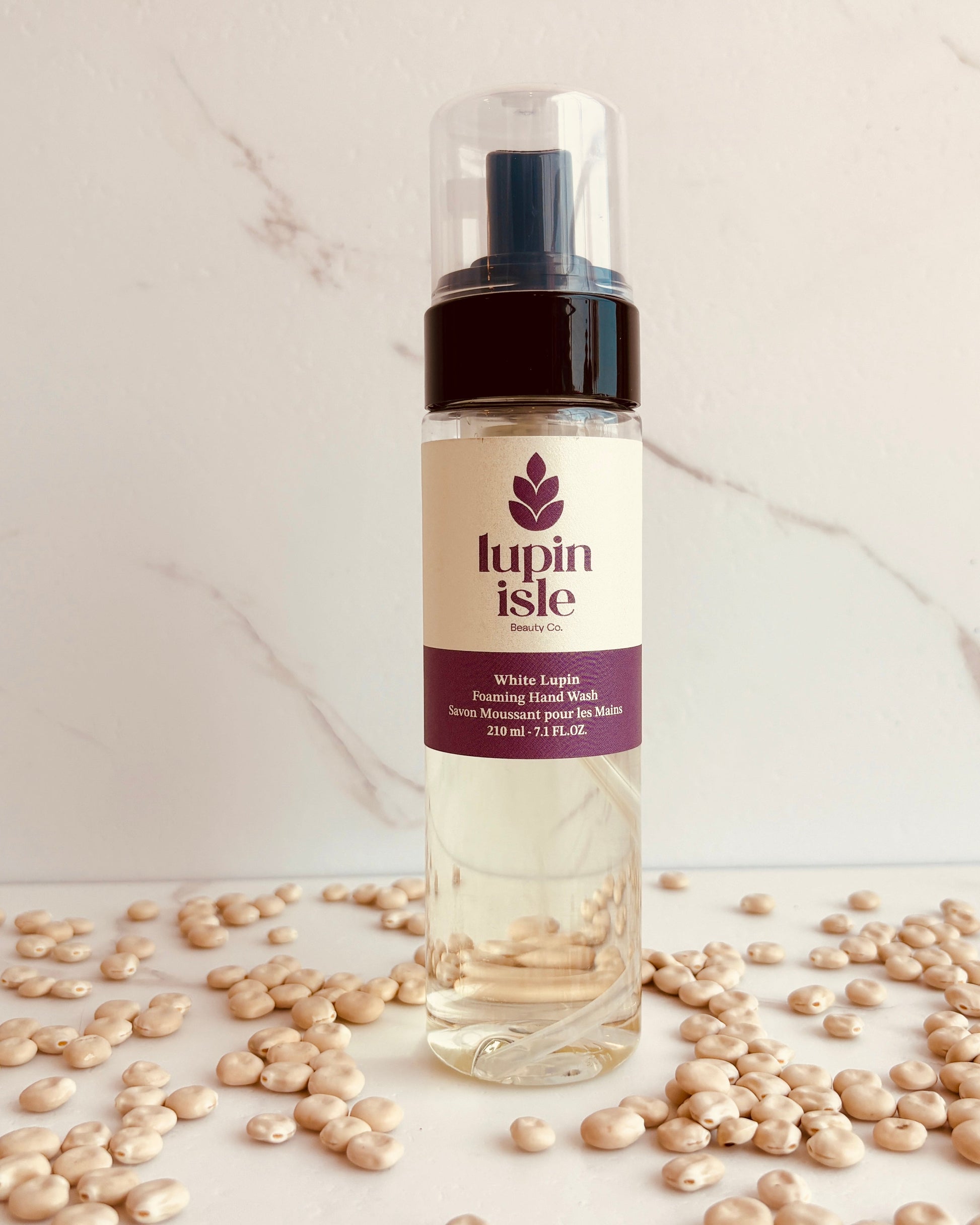 A bottle of Lupin Isle White Lupin Hair Foaming Hand Wash standing on a marble-like surface scattered with white lupin beans.