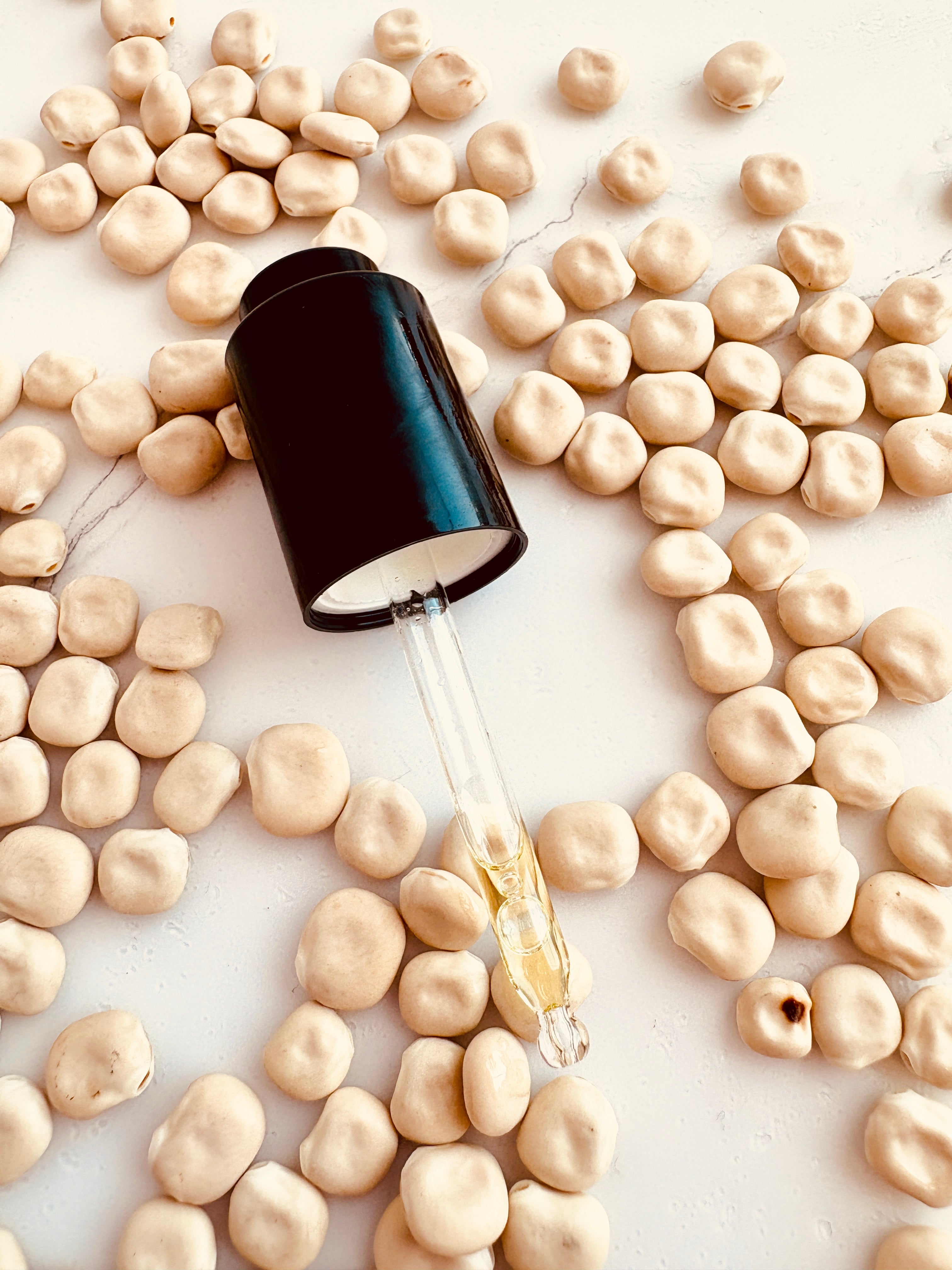 A clear glass dropper bottle with a black cap lies open on a white surface, surrounded by scattered lupin beans. A few drops of golden liquid are visible in the dropper.