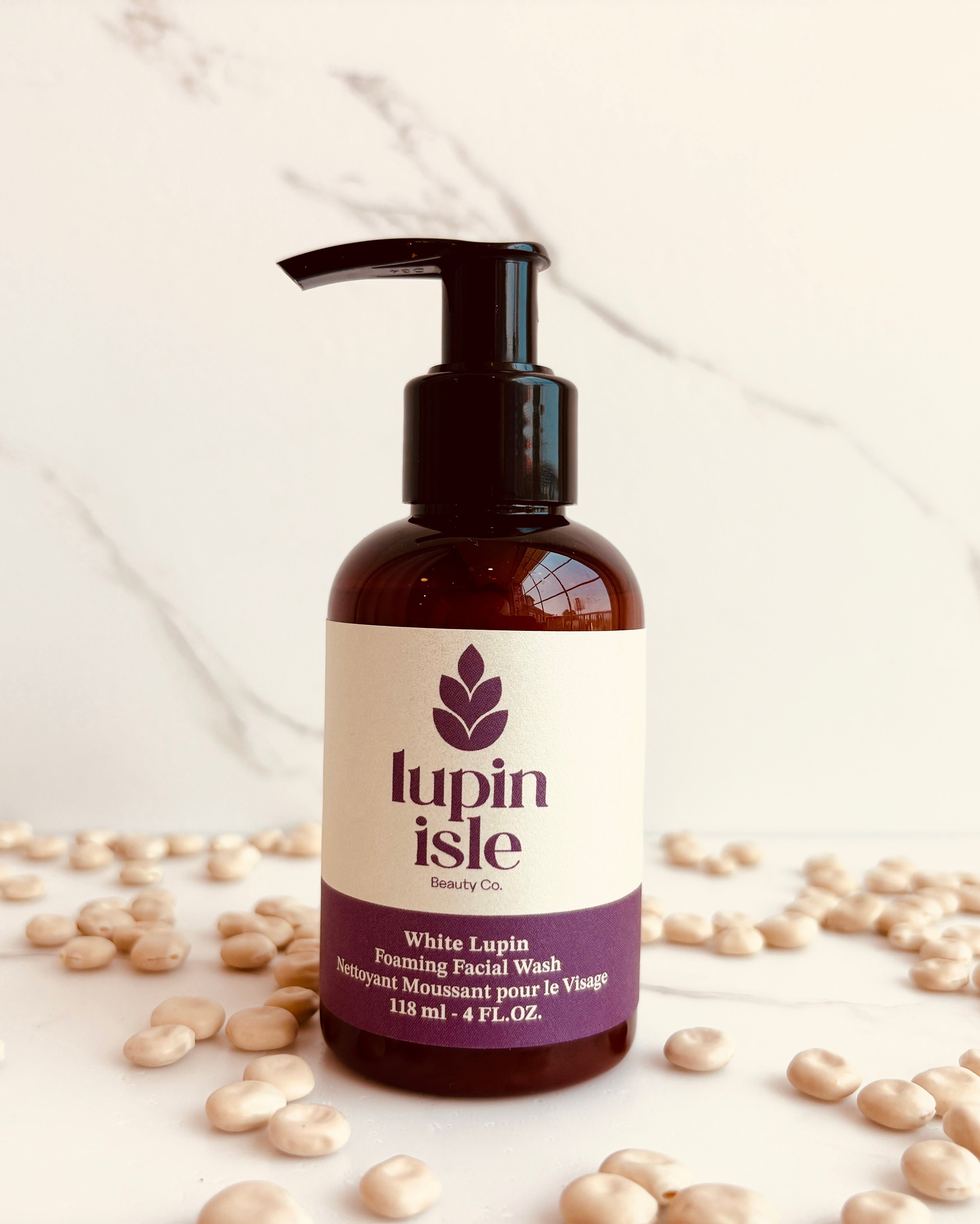 A bottle of Lupin Isle White Lupin Cleansing Facial Wash with a black pump dispenser, set against a white marble background and surrounded by scattered white lupin beans.