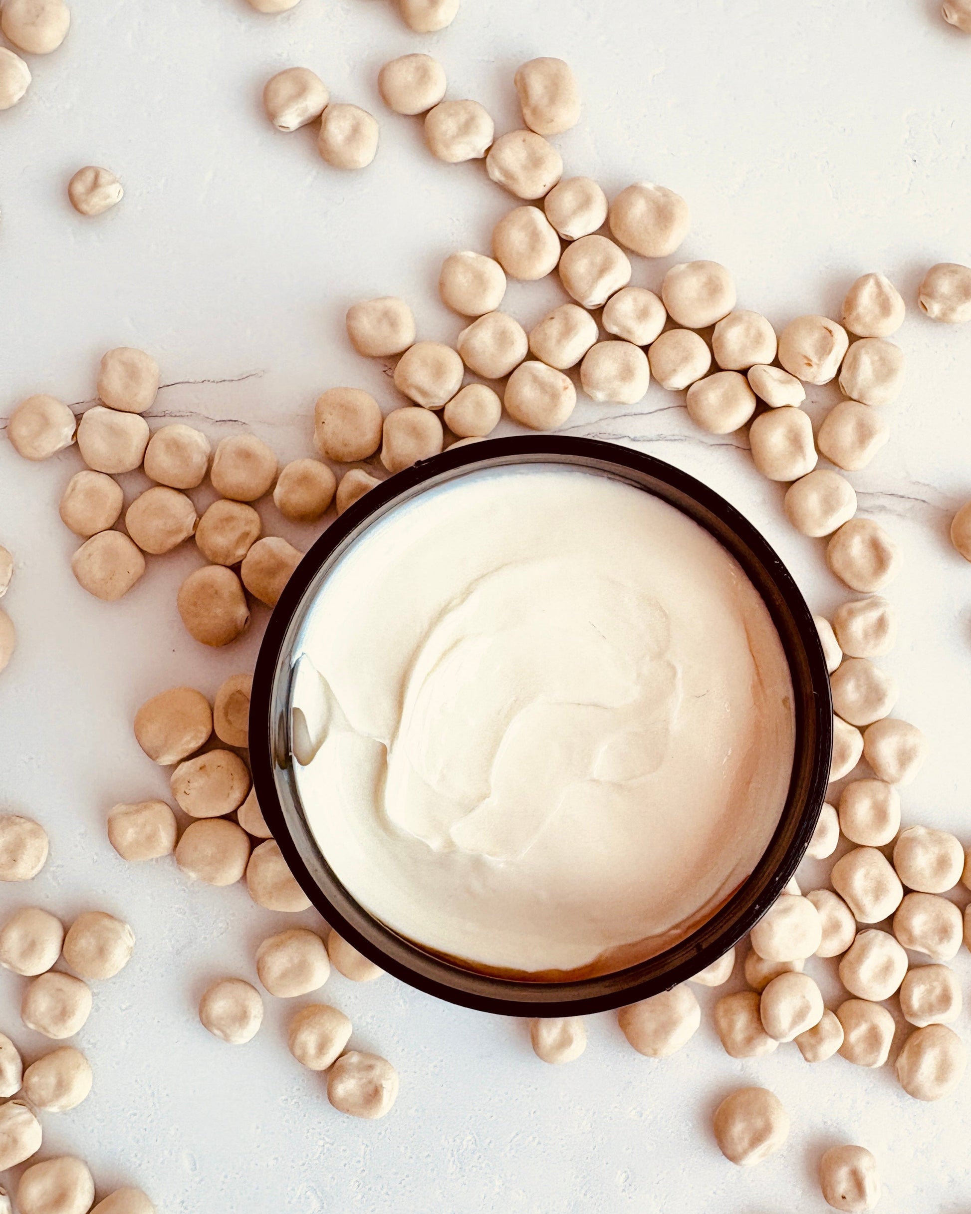 A jar of opened Lupin Isle Velvet Body Butter displaying the creamy texture, surrounded by scattered lupin seeds on a light neutral background.