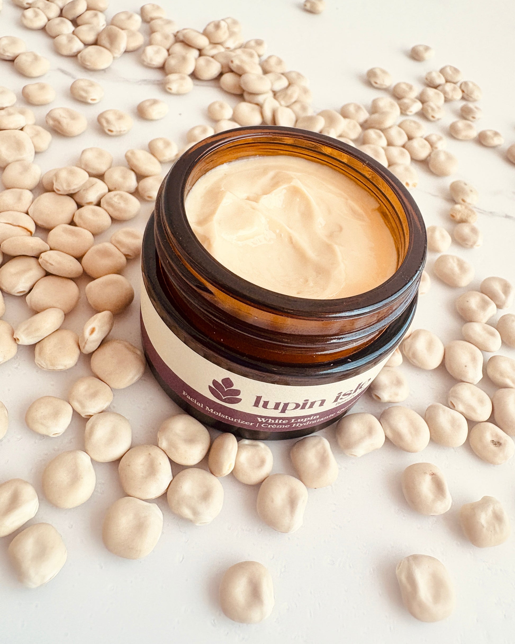 An open amber glass jar of Lupin Isle White Lupin Facial Moisturizer, surrounded by scattered lupin beans on a white surface.