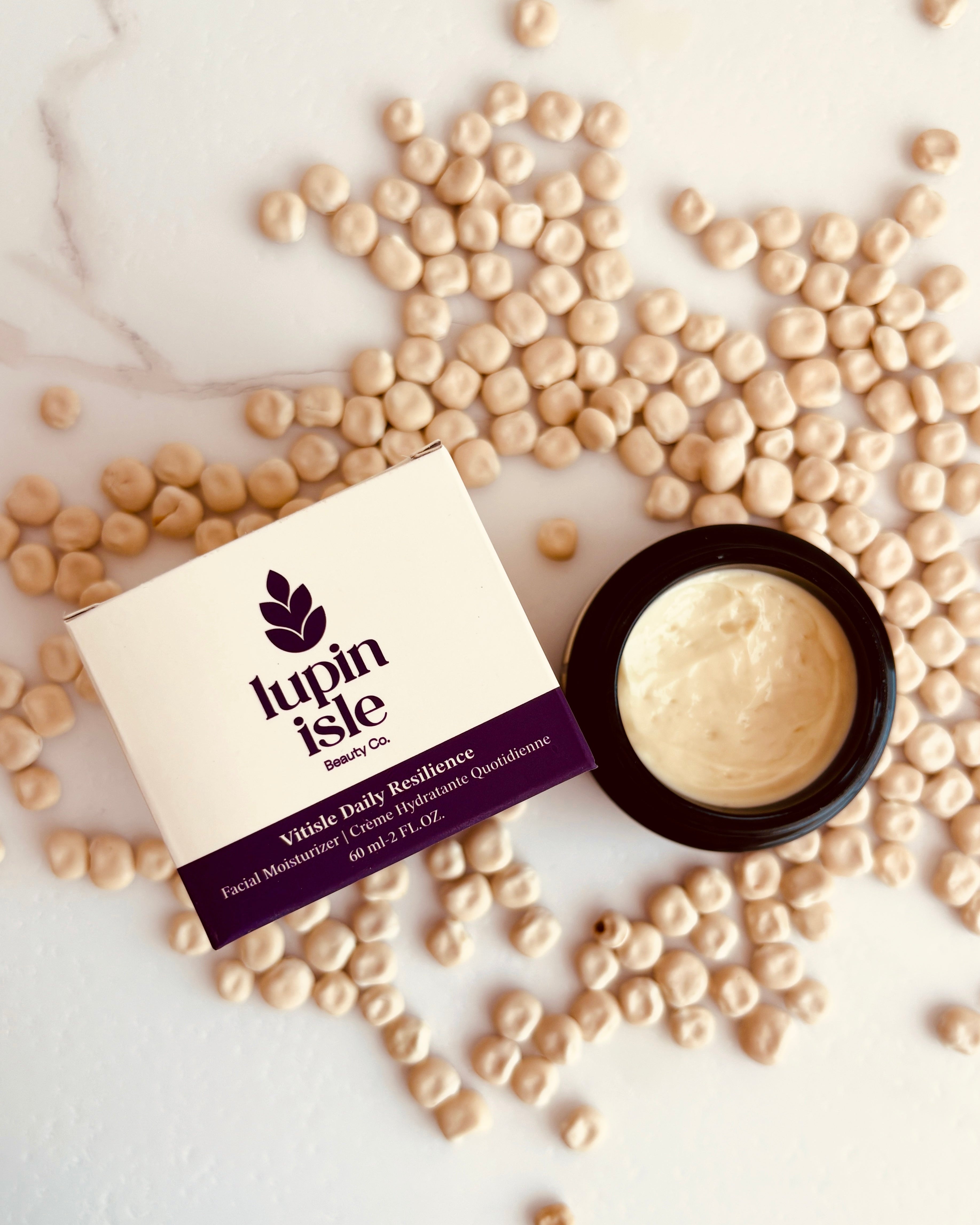 A flat-lay of Lupin Isle Vitale Daily Excellence facial moisturizer, showing the cream in an open black jar next to its cream-colored box, surrounded by scattered beige lupin seeds on a white marble background.