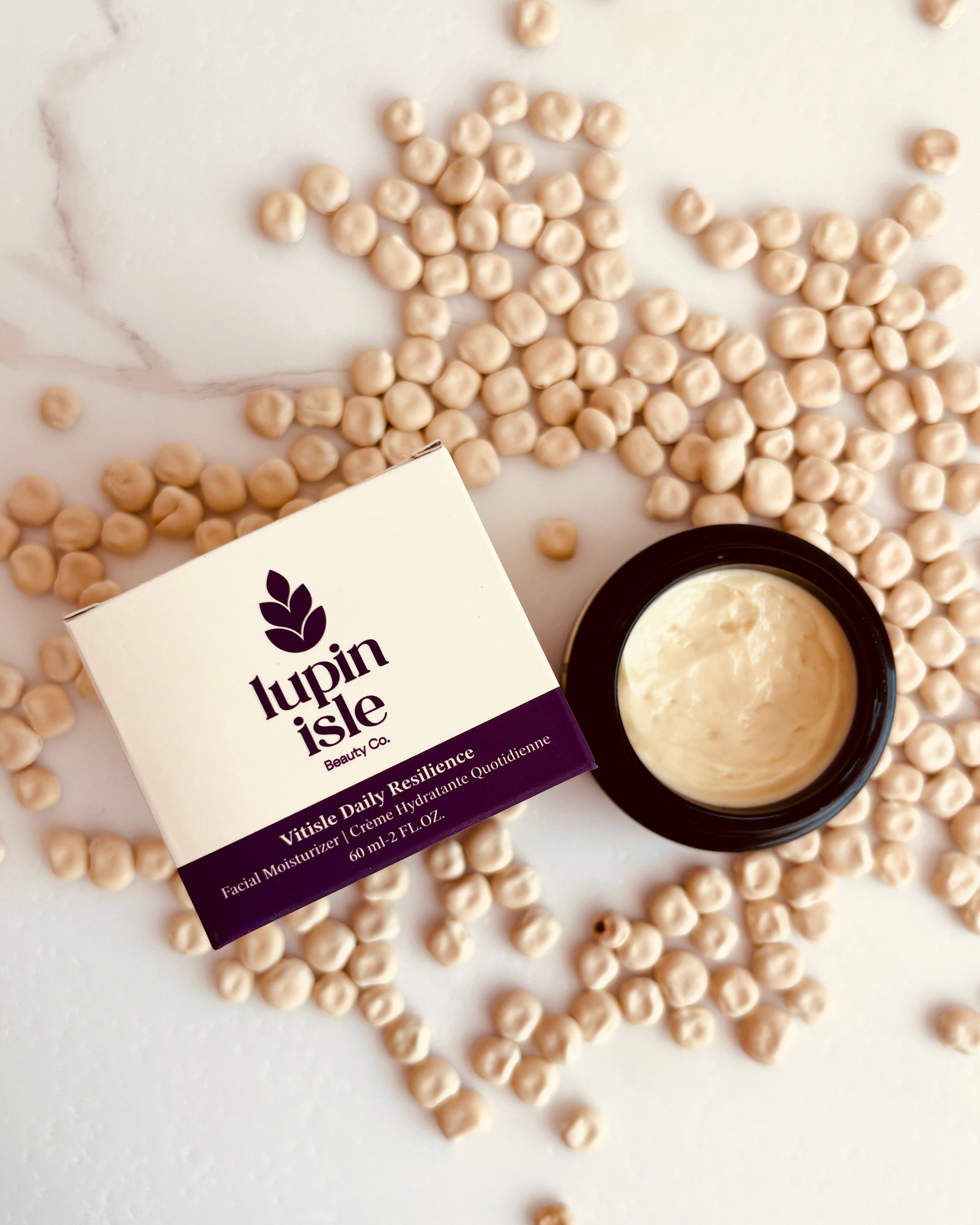 A flat-lay of Lupin Isle Vitale Daily Excellence facial moisturizer, showing the cream in an open black jar next to its cream-colored box, surrounded by scattered beige lupin seeds on a white marble background.