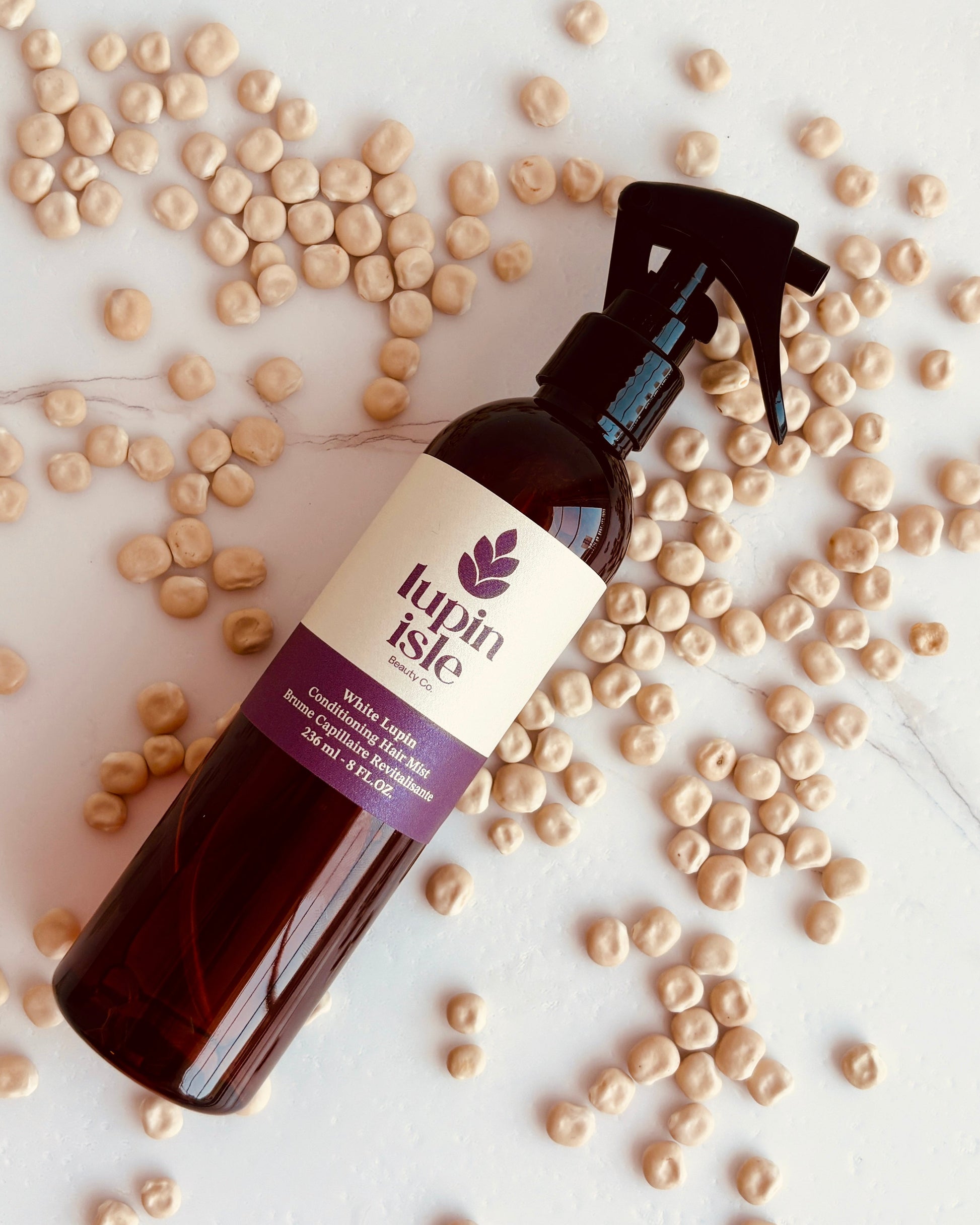 A bottle of Lupin Isle Conditioning Hair mist with a black spray top and cream label, standing on a marble surface surrounded by scattered lupin beans.