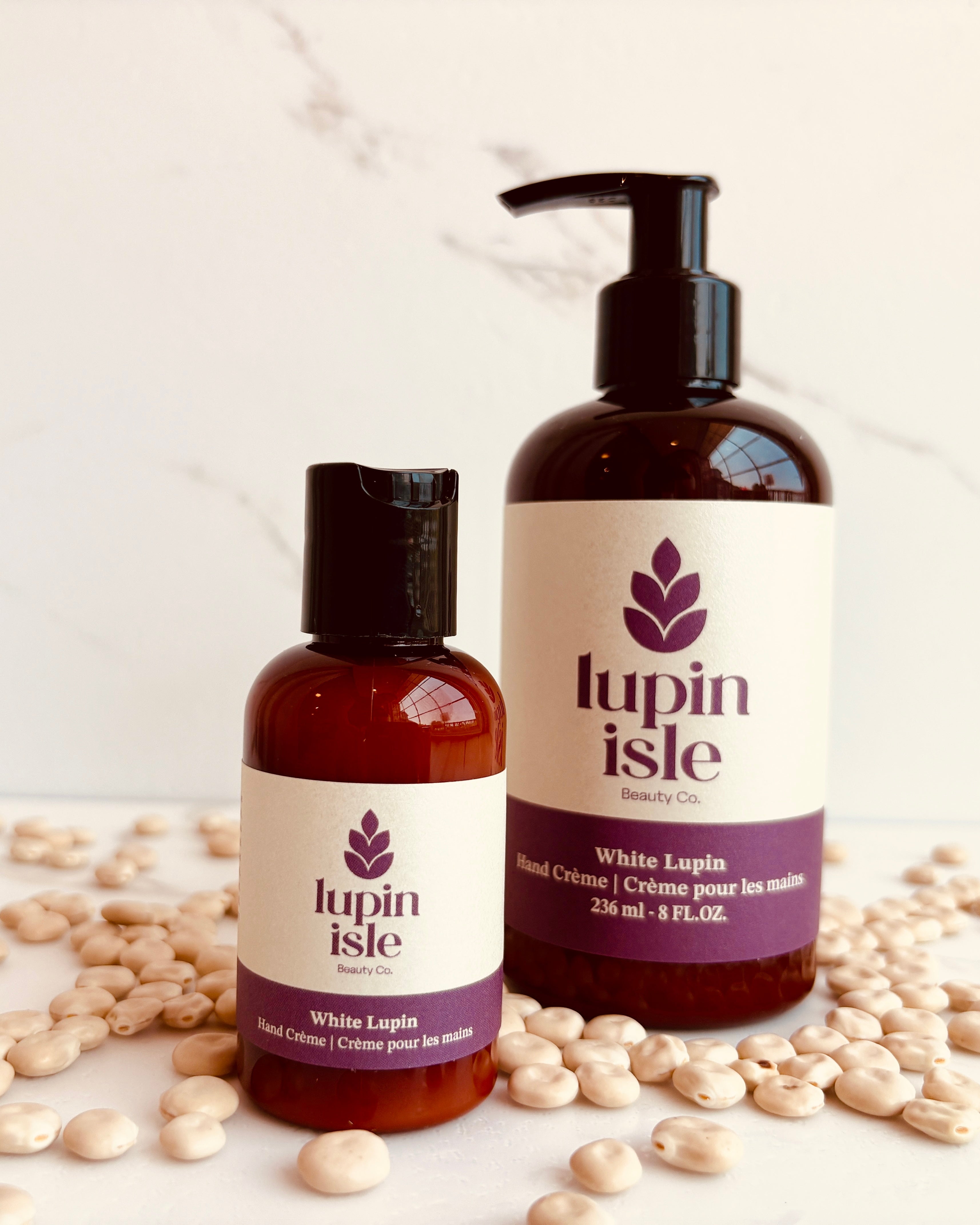 Two amber bottles of Lupin Isle White Lupin hand cream arranged on a white marble surface with scattered white lupin beans. The larger pump bottle stands on the right, and the smaller flip-top bottle stands on the left.