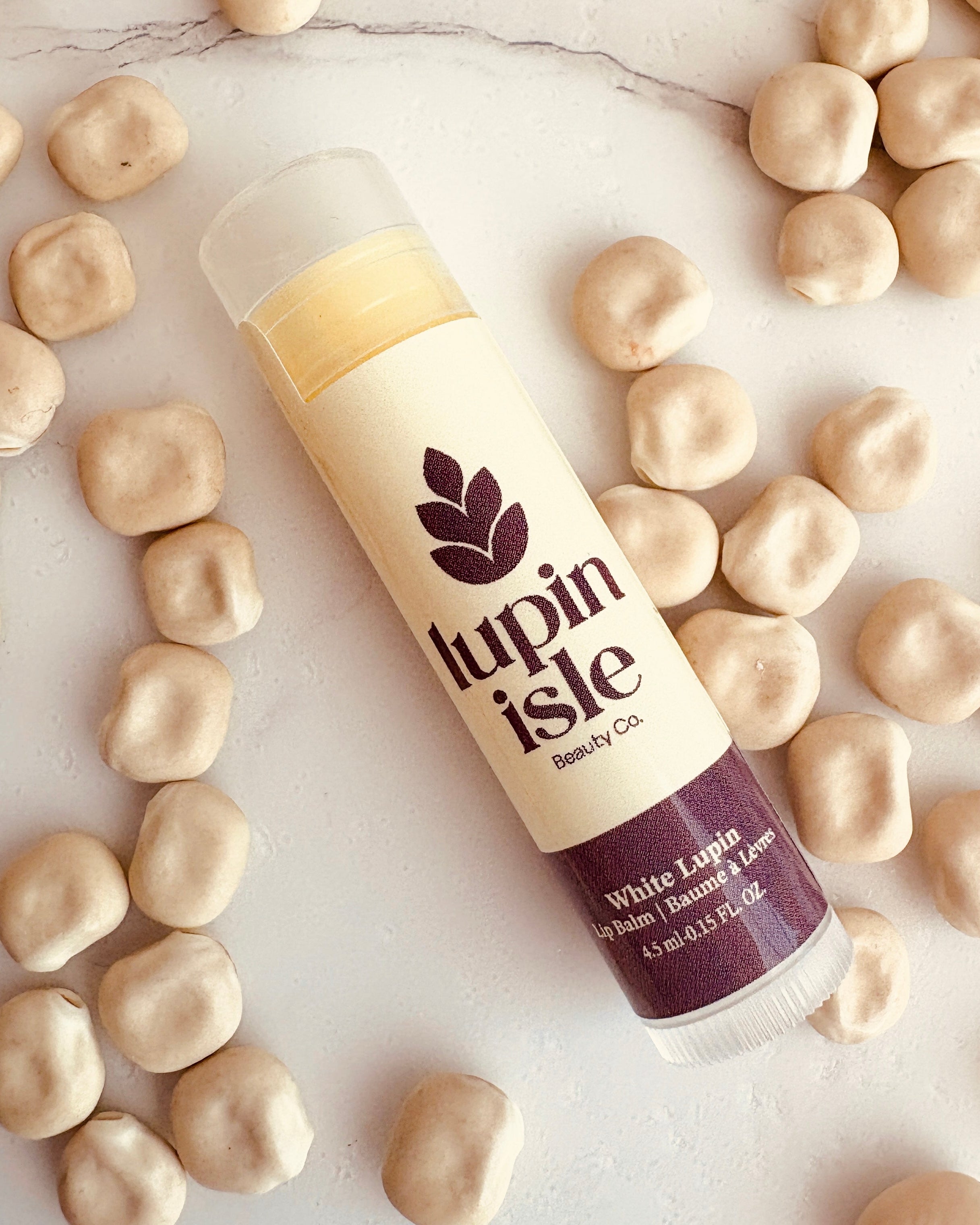 A tube of Lupin Isle White Lip Balm is shown diagonally on a white marble surface, surrounded by scattered beige lupin beans. The product features a cream-colored label with dark purple text and logo, and a dark purple cap.