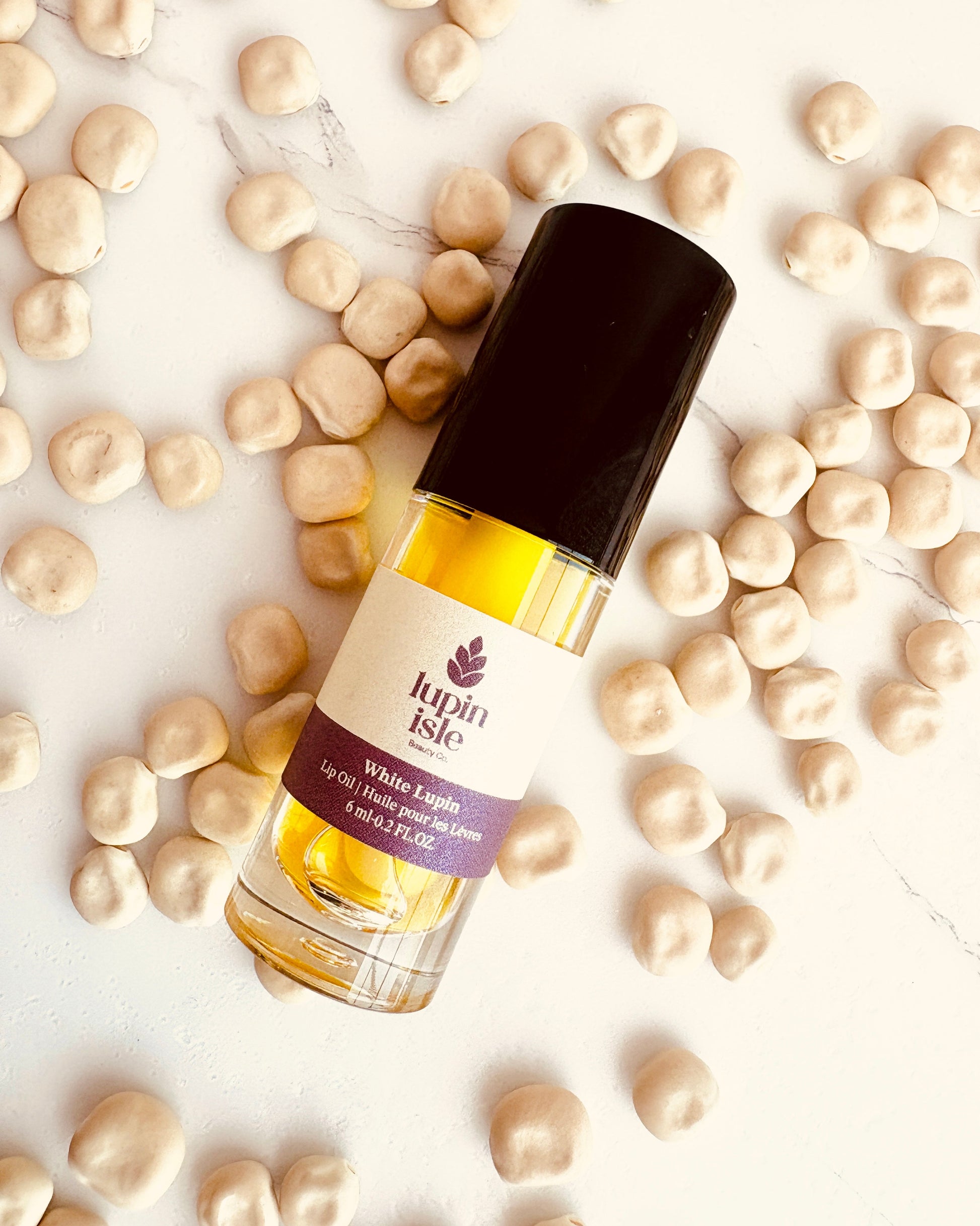 A bottle of Lupin Isle White Lupin lip oil with a black cap, surrounded by scattered beige lupin seeds on a white marble background.