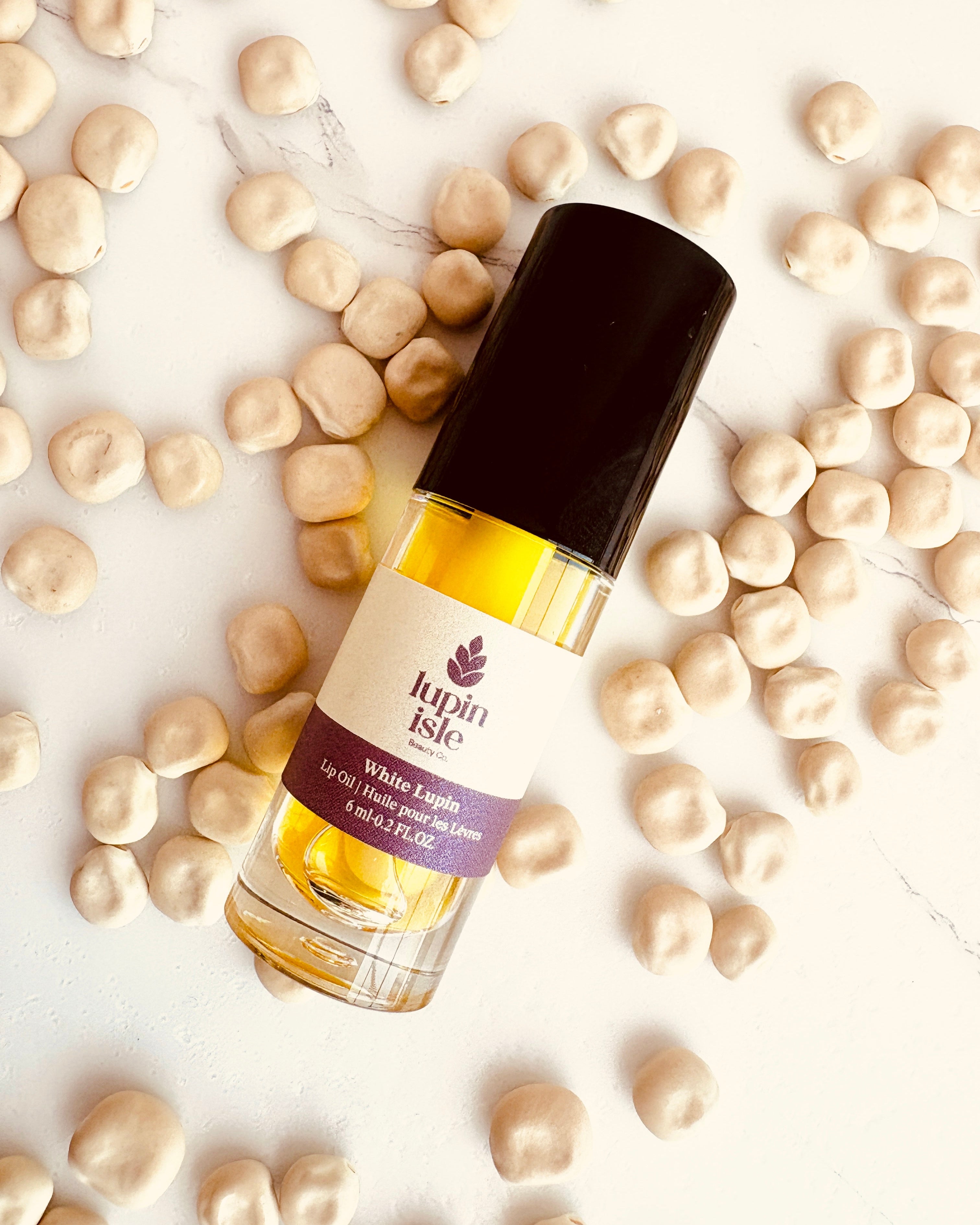 A bottle of Lupin Isle White Lupin lip oil with a black cap, surrounded by scattered beige lupin seeds on a white marble background.