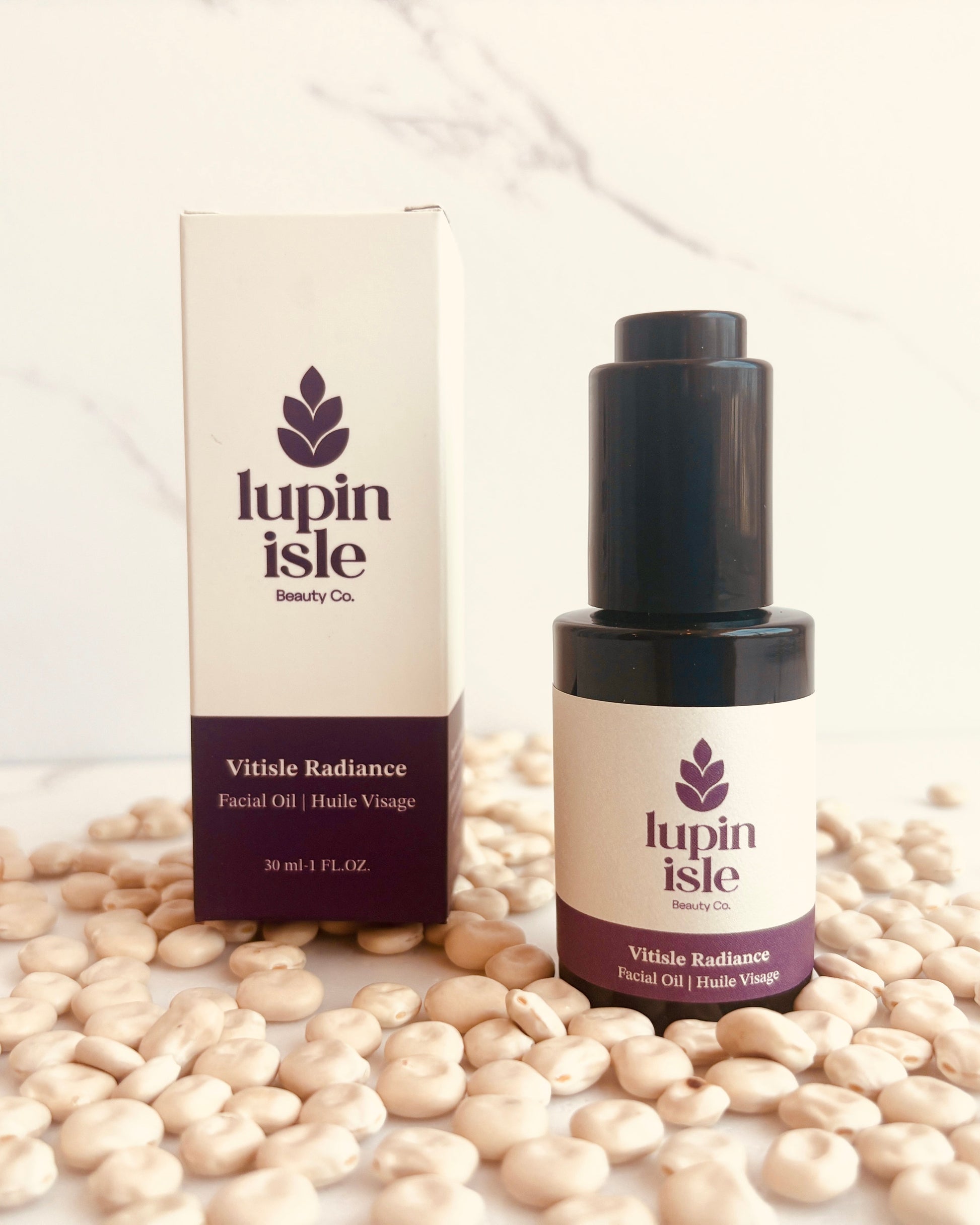 A bottle and box of Lupin Isle Vitisse Radiance Facial Oil arranged on a marble surface scattered with white beans.