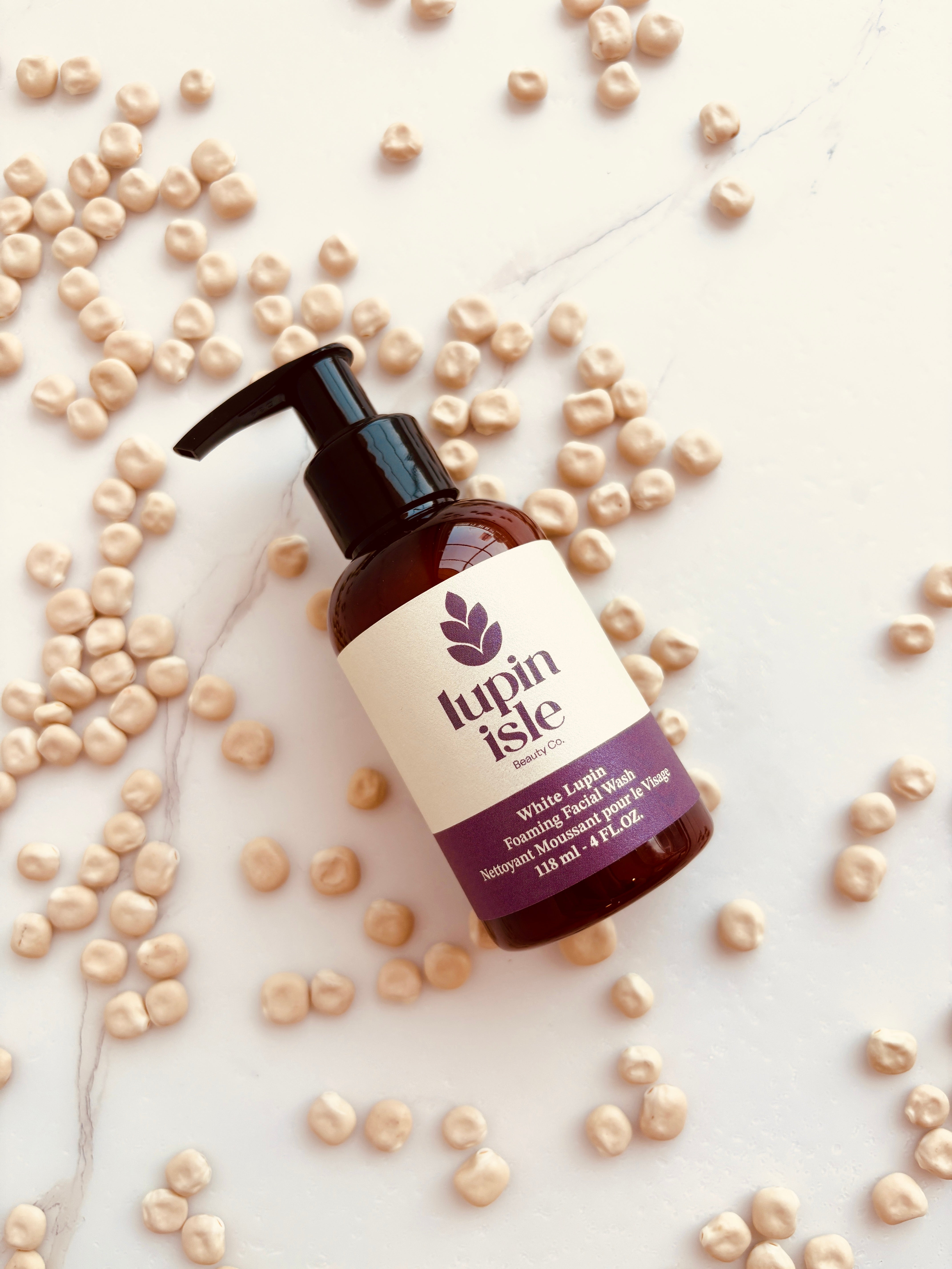 A bottle of Lupin Isle Foot Washing Solution with a black pump dispenser, placed on a white marble surface surrounded by scattered lupin beans.