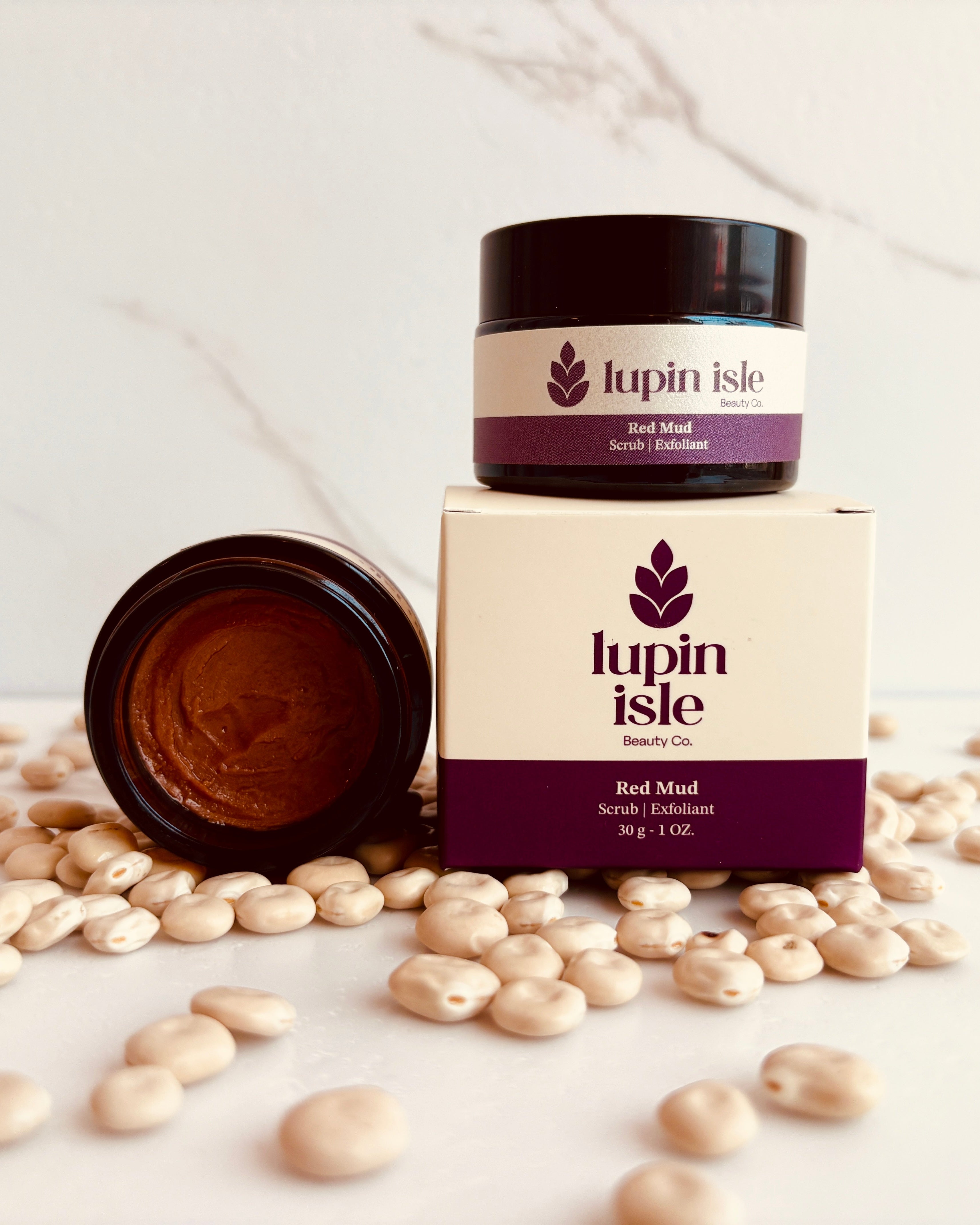 A jar of Lupin Isle Red Mud Scrub stands upright on top of its cream-colored and purple box, with the jar lid lying beside it. The scene is styled with scattered lupin beans on a light marble surface and background.