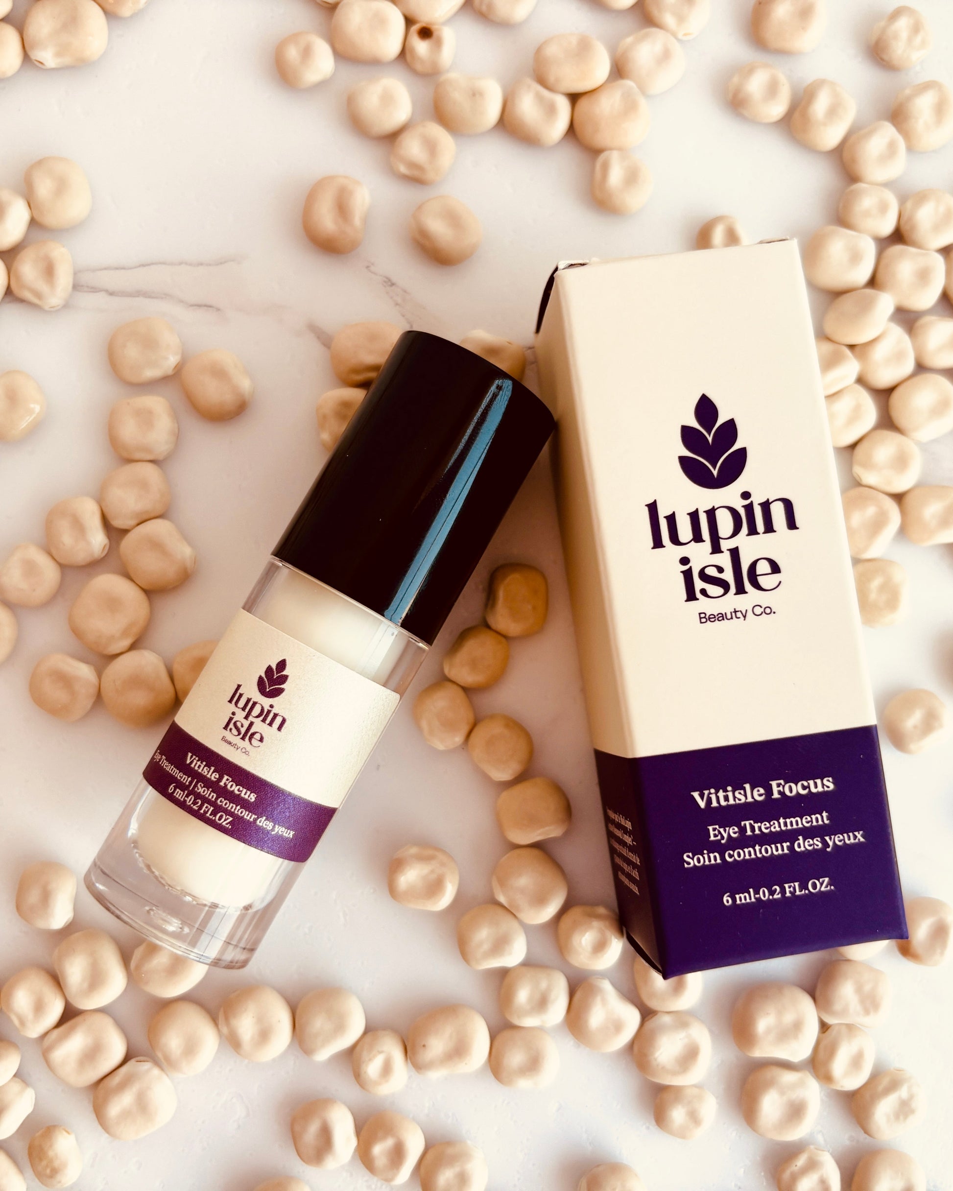 A bottle of lupin Isle Vitide Focus Eye Treatment and its matching box, styled on a white marble surface scattered with beige lupin seeds.
