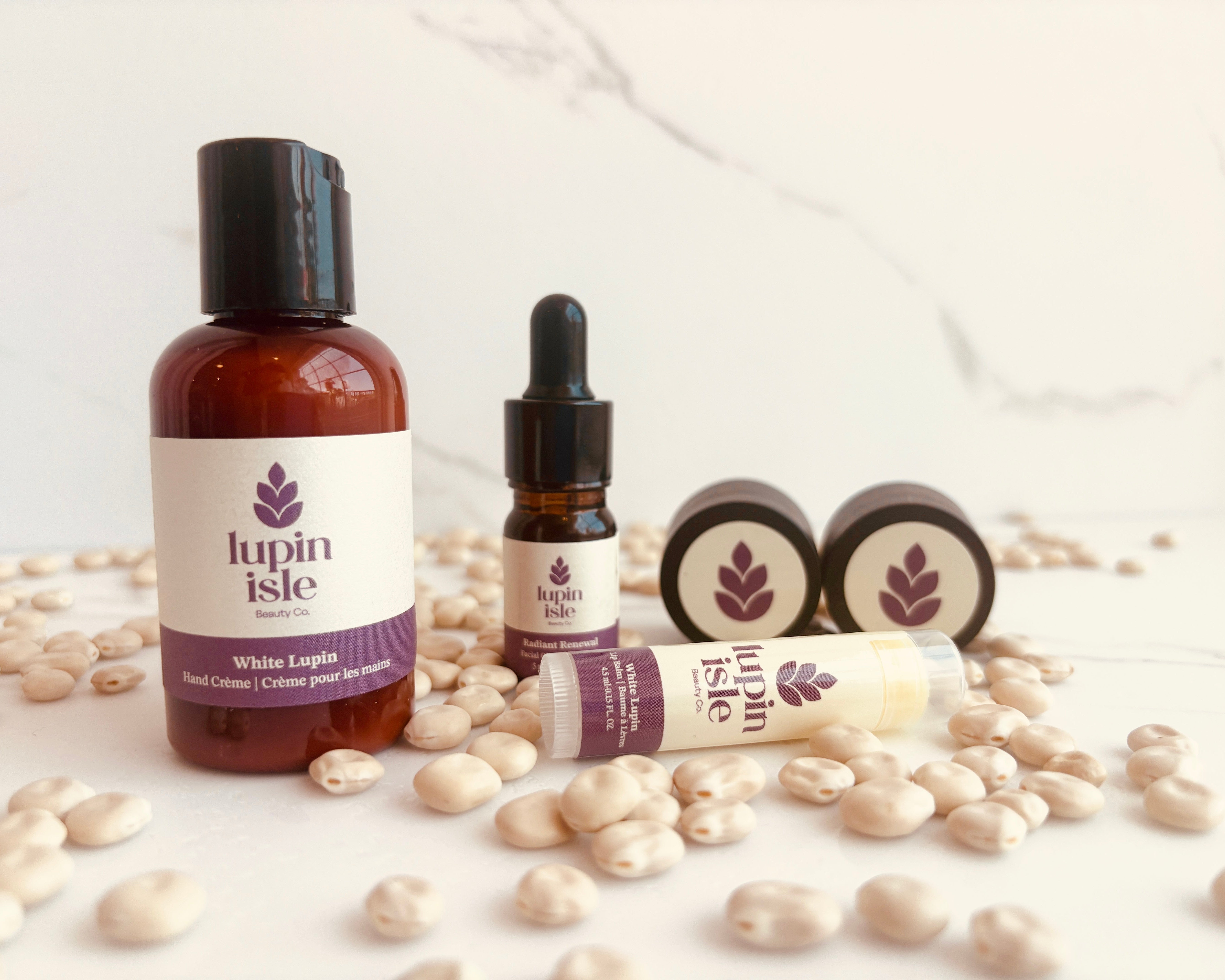 A set of Lupin Isle skincare products arranged on a light marble surface scattered with lupin beans. The set includes a brown bottle of White Lupin Hand Crème, Radiant Renewal Facial Oil, Red Mud Scrub, White lupin Facial Moisturizer and a lip balm.