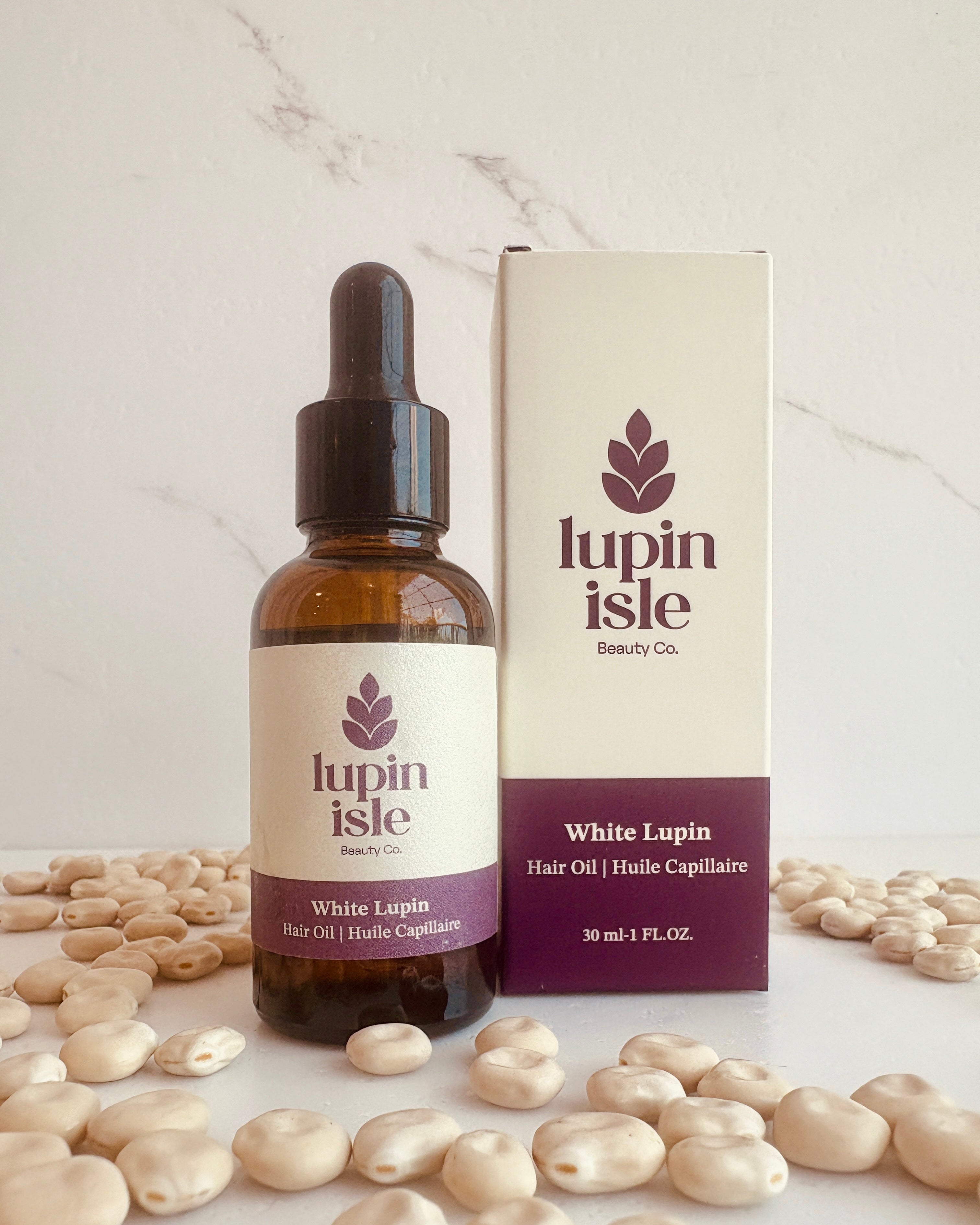 A bottle of Lupin Isle Beauty Co. White Lupin Hair Oil next to its cream and purple packaging box, styled with white lupin beans on a light marble surface.