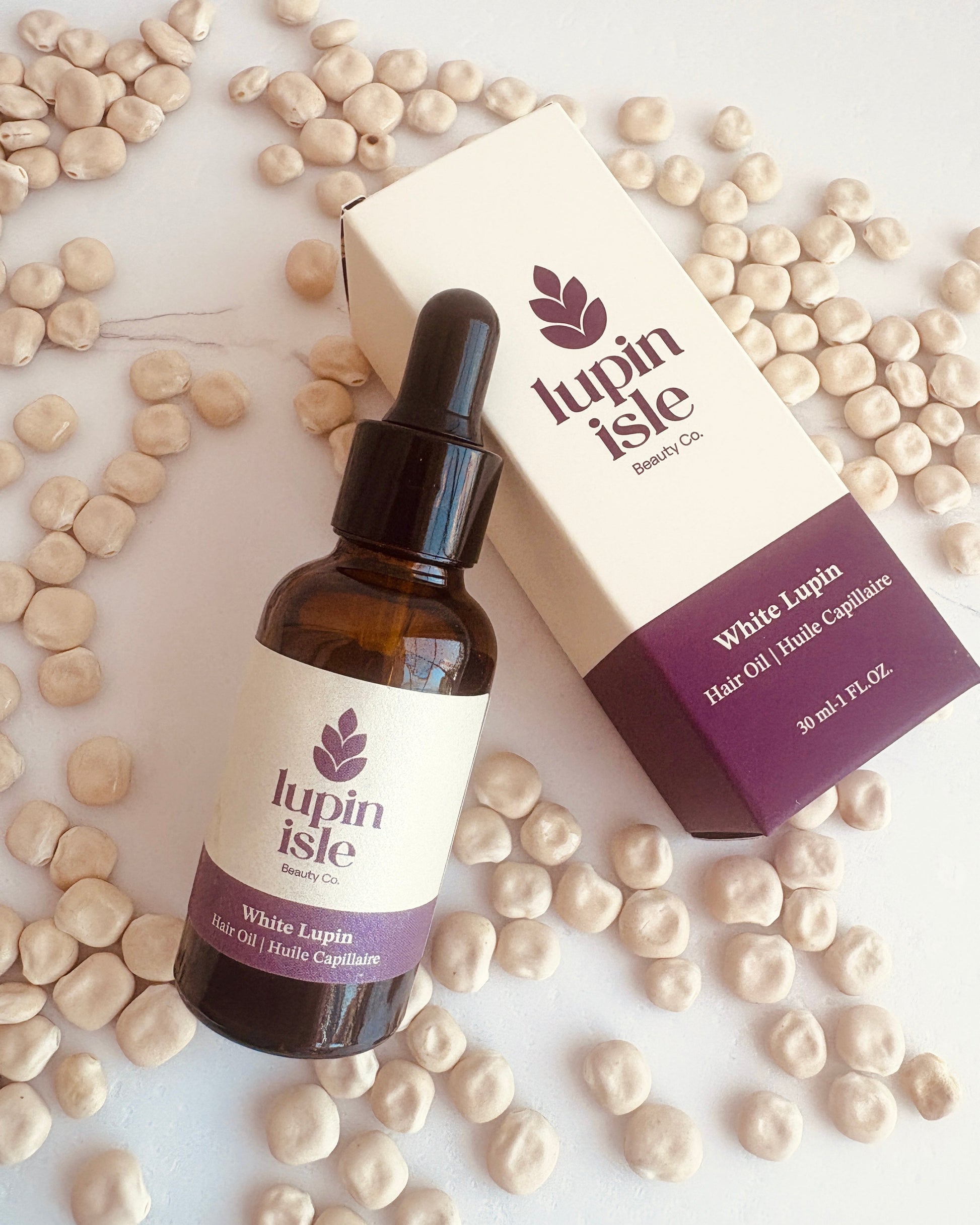 A bottle of lupin Isle White Lupin Hair Oil next to its product box, styled on a white surface with scattered beige lupin seeds.